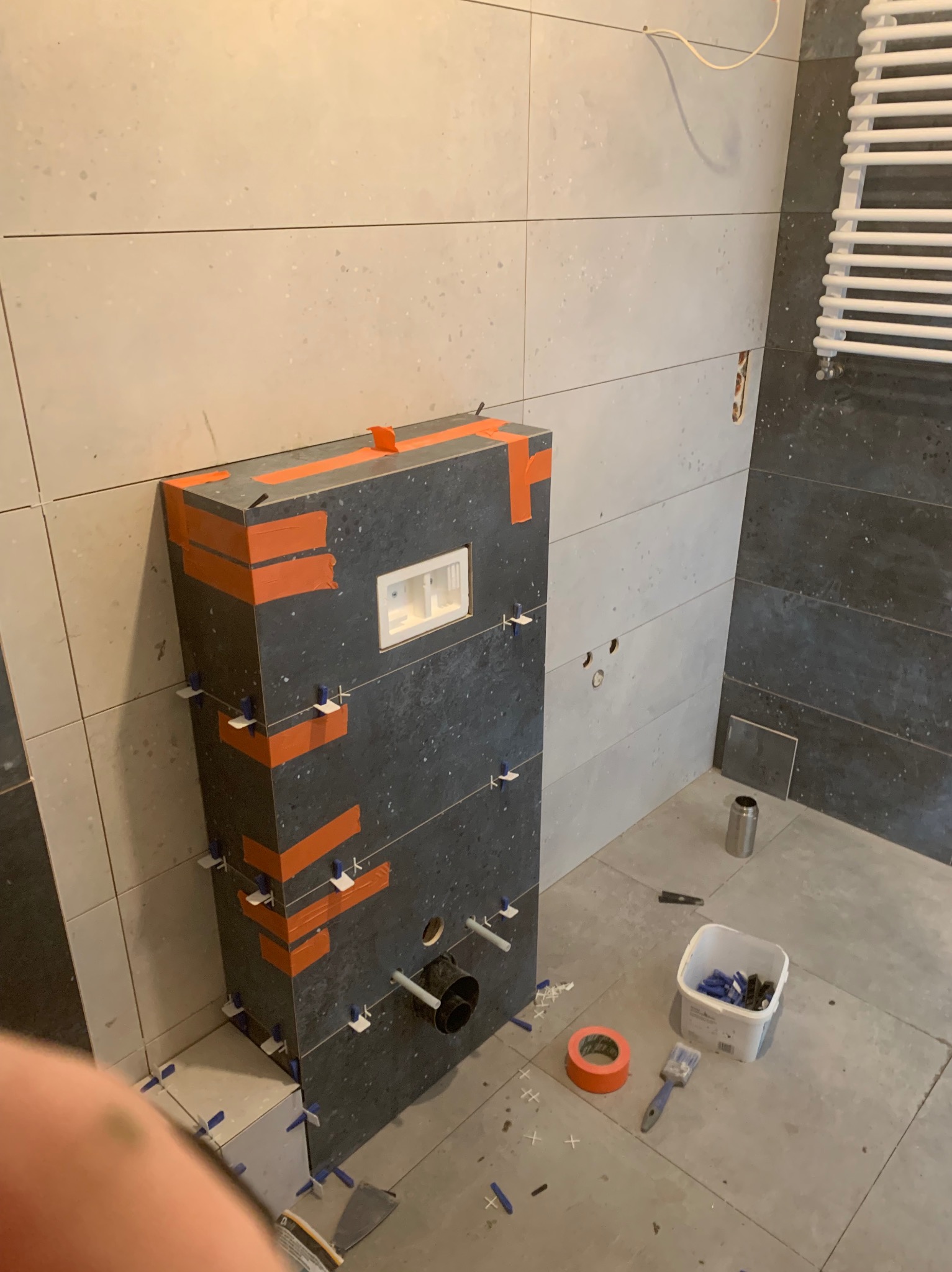 Montaż płytek na obudowie stelaża WC z użyciem pomarańczowej taśmy i klipsów poziomujących. Widoczne narzędzia glazurnicze i fragment grzejnika.