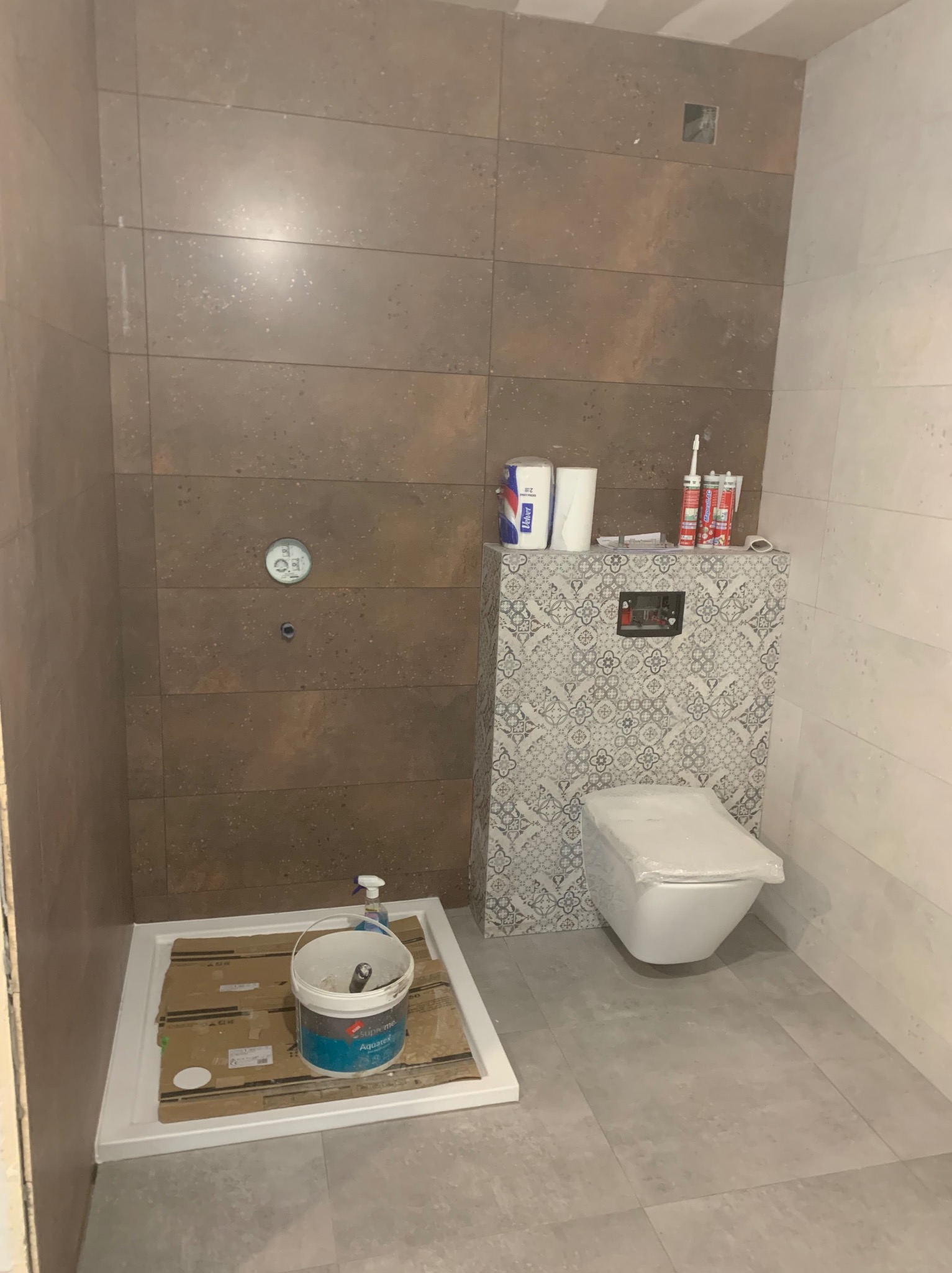 Remont łazienki: brązowe płytki ścienne, podwieszana toaleta z wzorzystą obudową, brodzik i wiadro z farbą. Widoczne materiały budowlane i narzędzia.