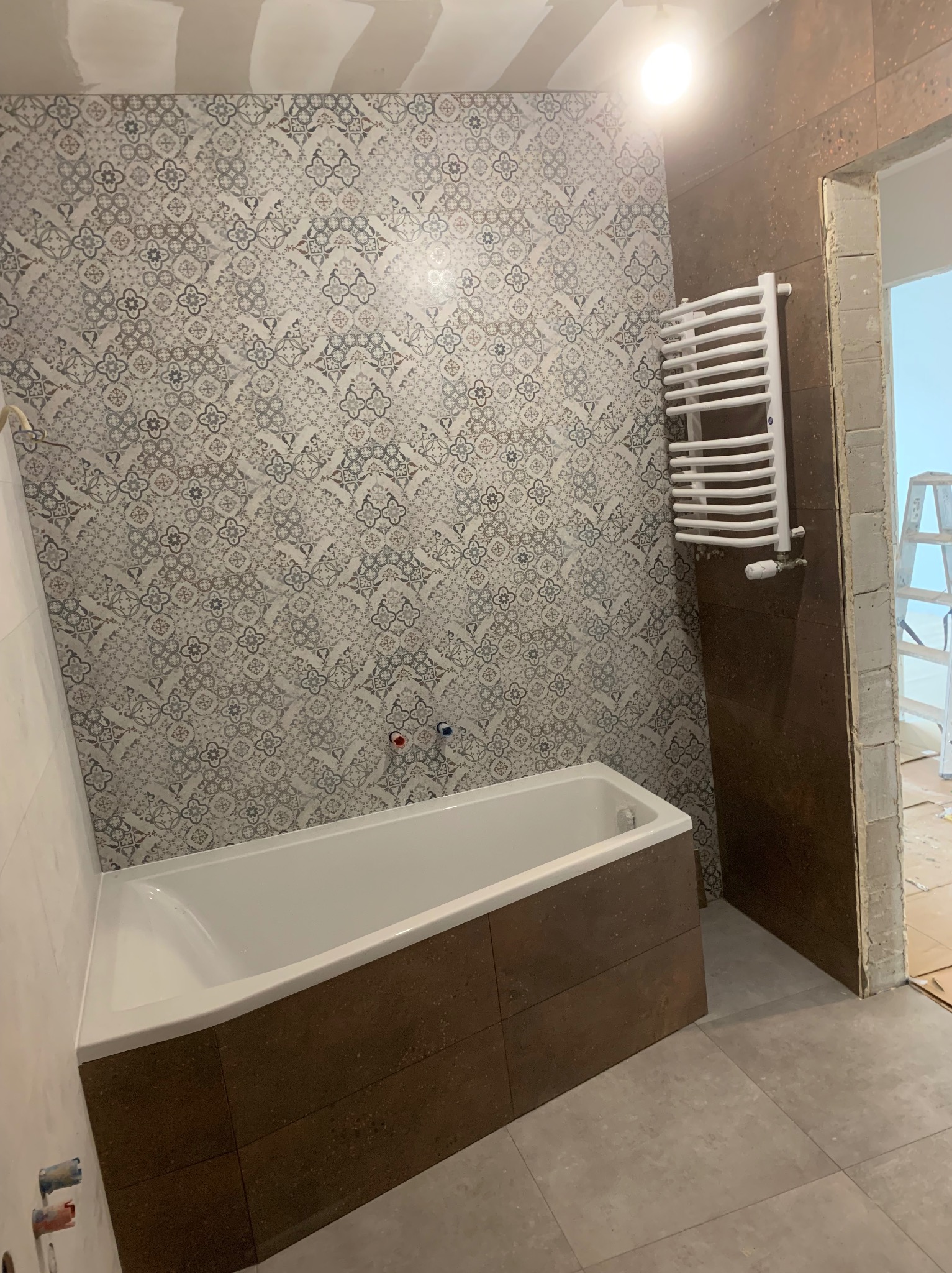 Wykończona łazienka z wanną obudowaną brązowymi płytkami, ozdobną mozaiką na ścianie i białym grzejnikiem drabinkowym. Widoczne surowe wykończenie sufitu i futryny.