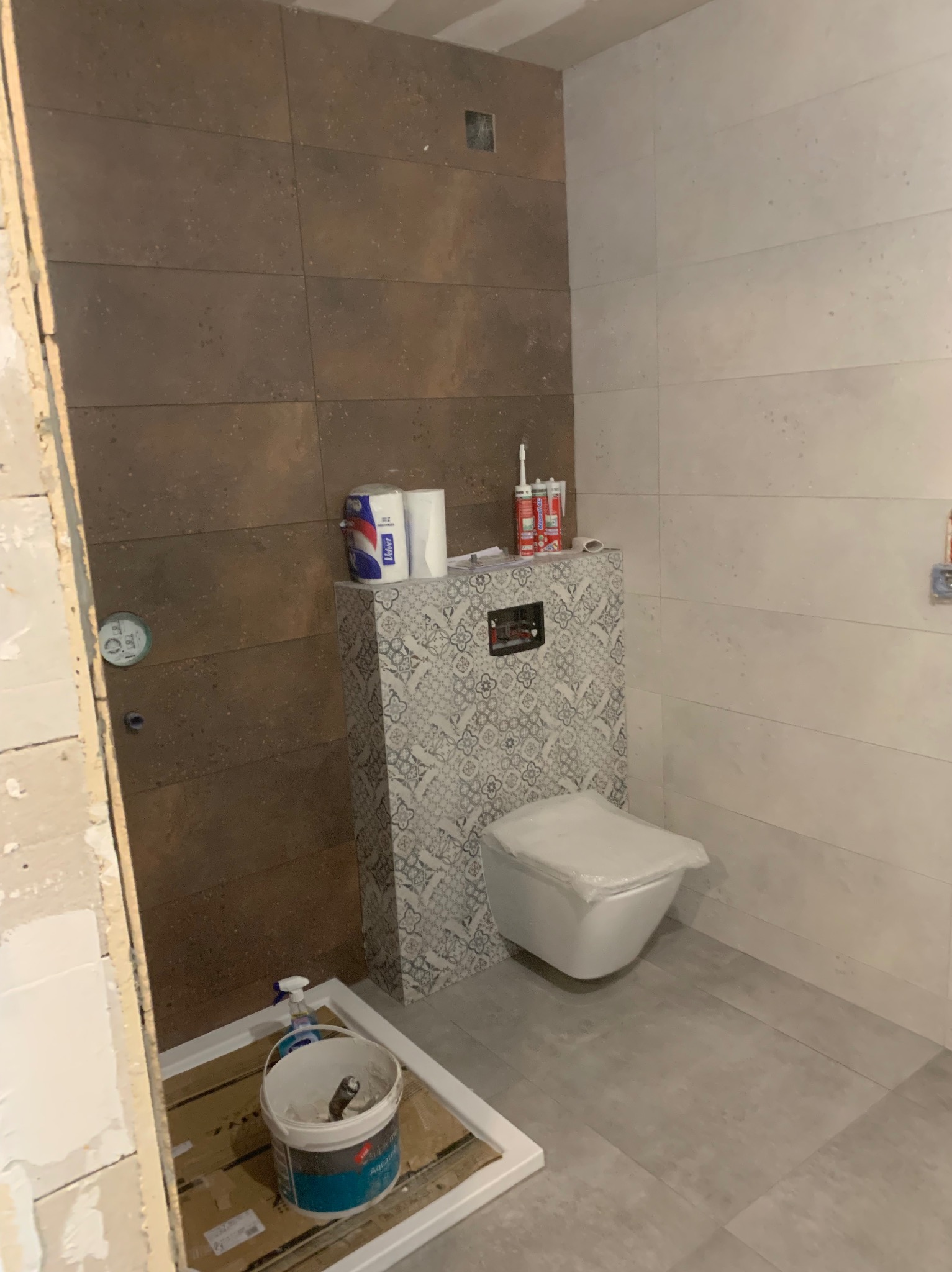 Remont łazienki w Zielonej Górze: brązowe i białe płytki na ścianach, podwieszana toaleta z wzorzystą obudową. Wiadro z narzędziami na podłodze.