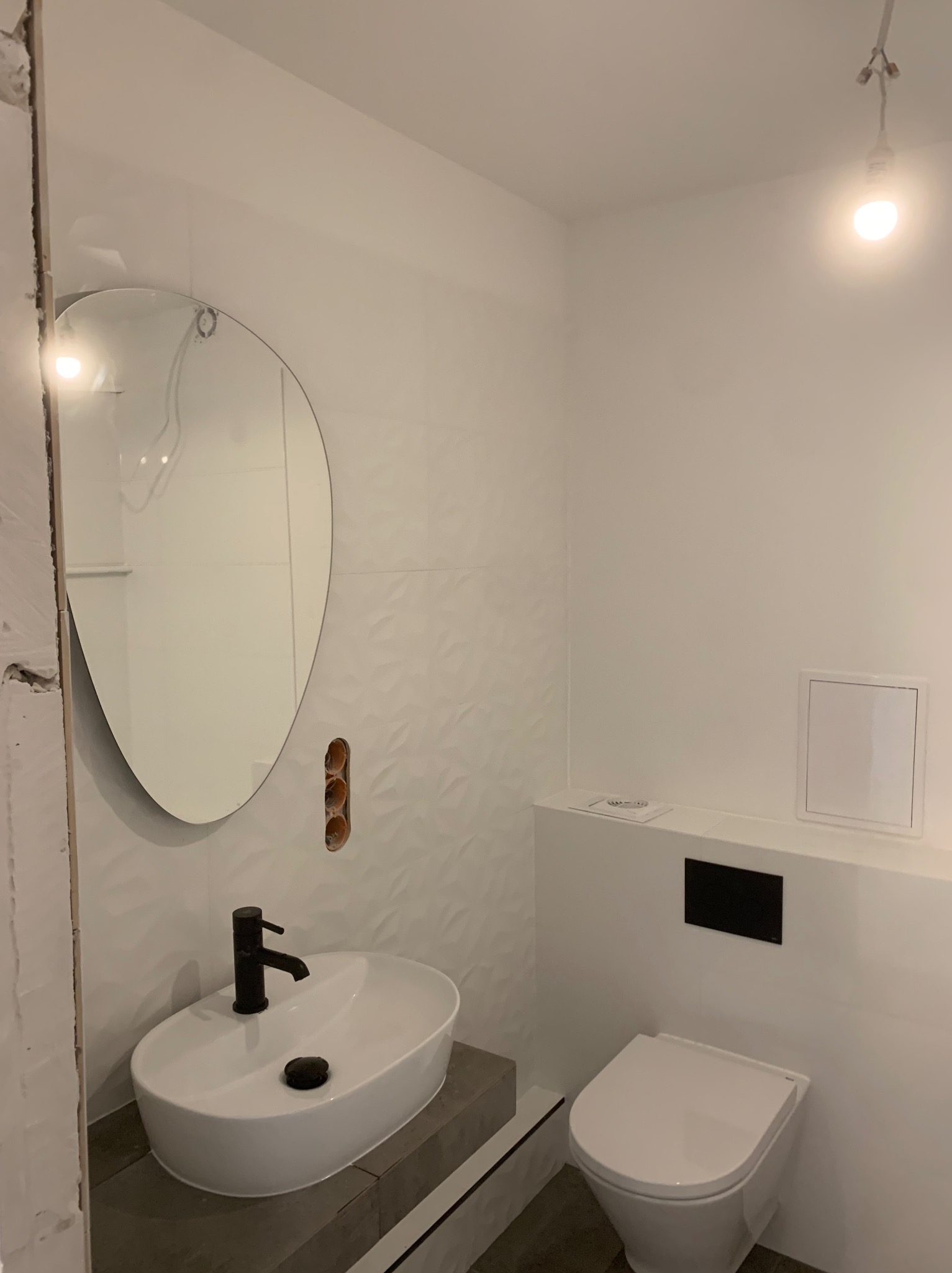 Biała łazienka z owalnym lustrem, umywalką nablatową i podwieszaną toaletą. Na ścianach białe płytki z subtelnym wzorem. Widoczne niedokończone wykończenie przy lustrze.