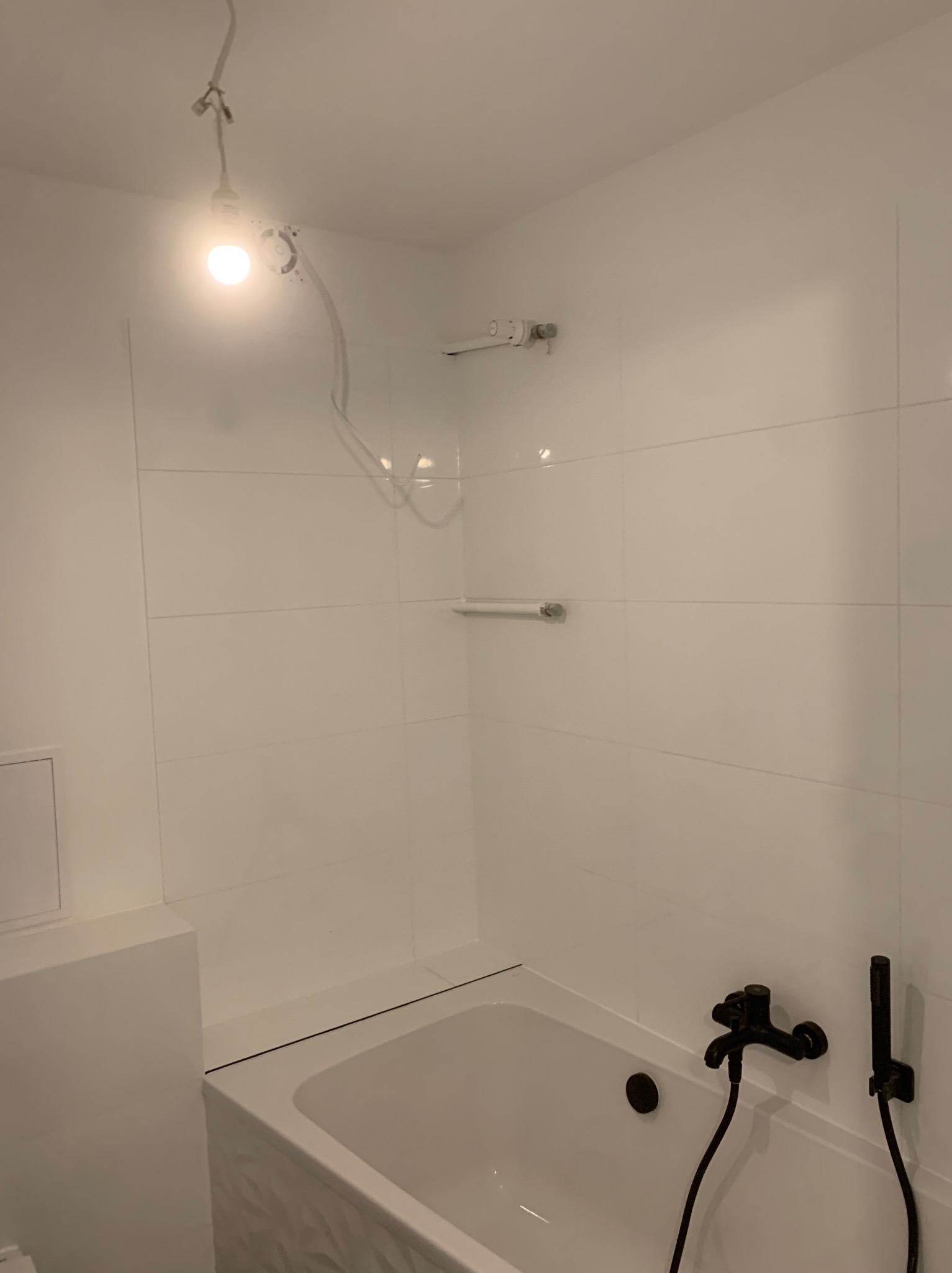 Biała łazienka z wanną i czarną armaturą. Ściany wyłożone białymi, prostokątnymi płytkami. Widoczne oświetlenie sufitowe i akcesoria łazienkowe.