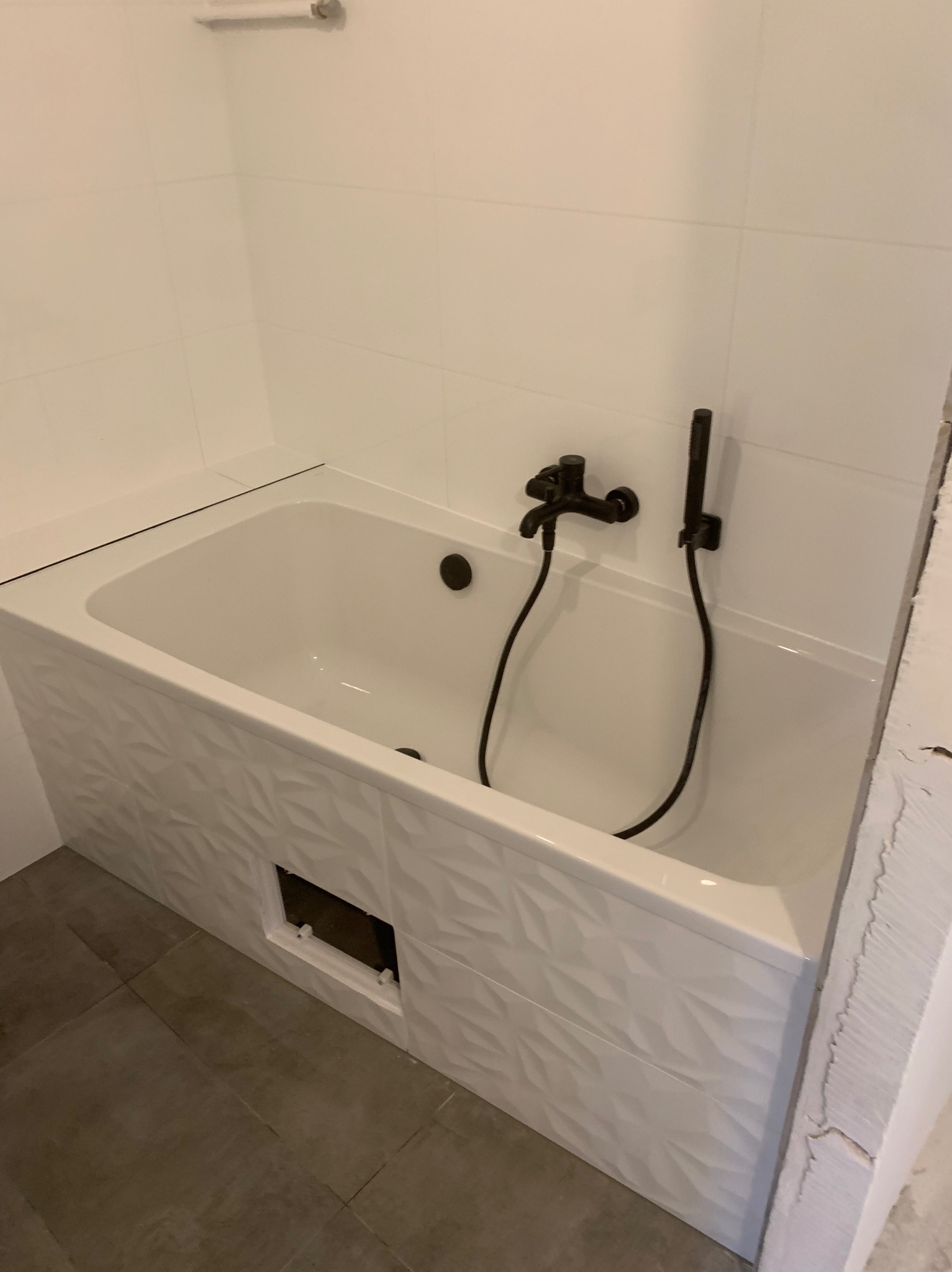 Biała wanna obudowana płytkami z geometrycznym wzorem, czarna bateria, szare płytki podłogowe. Widoczny otwór rewizyjny. Minimalistyczne wykończenie łazienki.