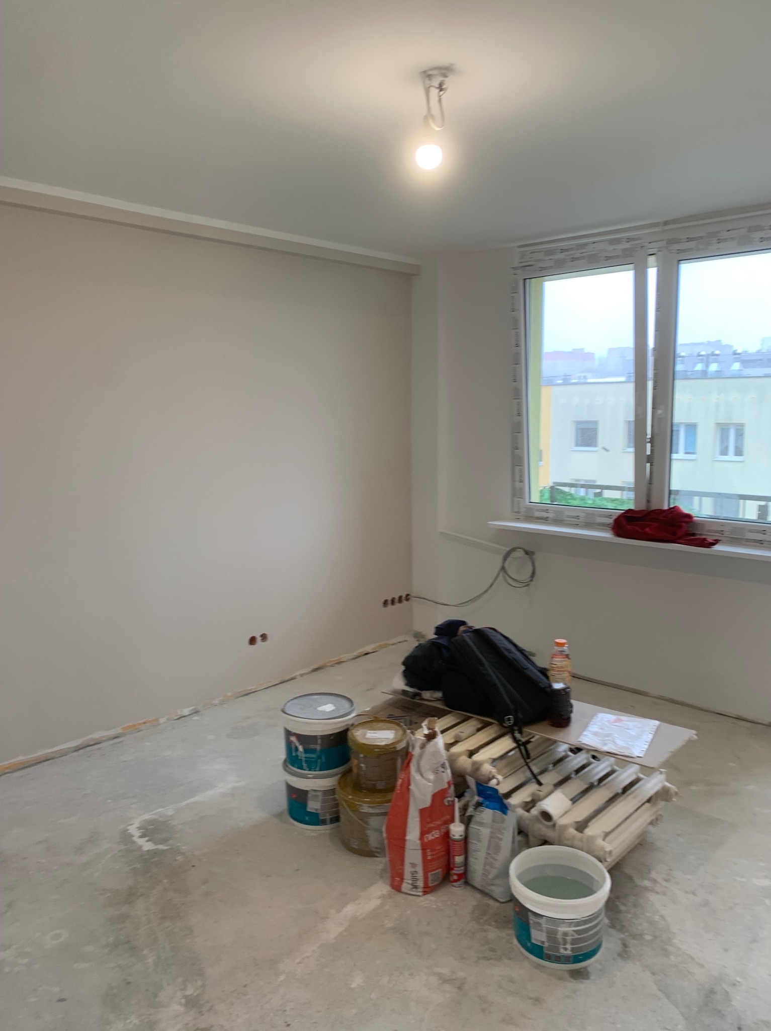 Wnętrze pokoju w trakcie malowania: świeżo pomalowane ściany w odcieniach bieli, widoczne puszki z farbą, narzędzia malarskie i grzejnik przygotowany do malowania.