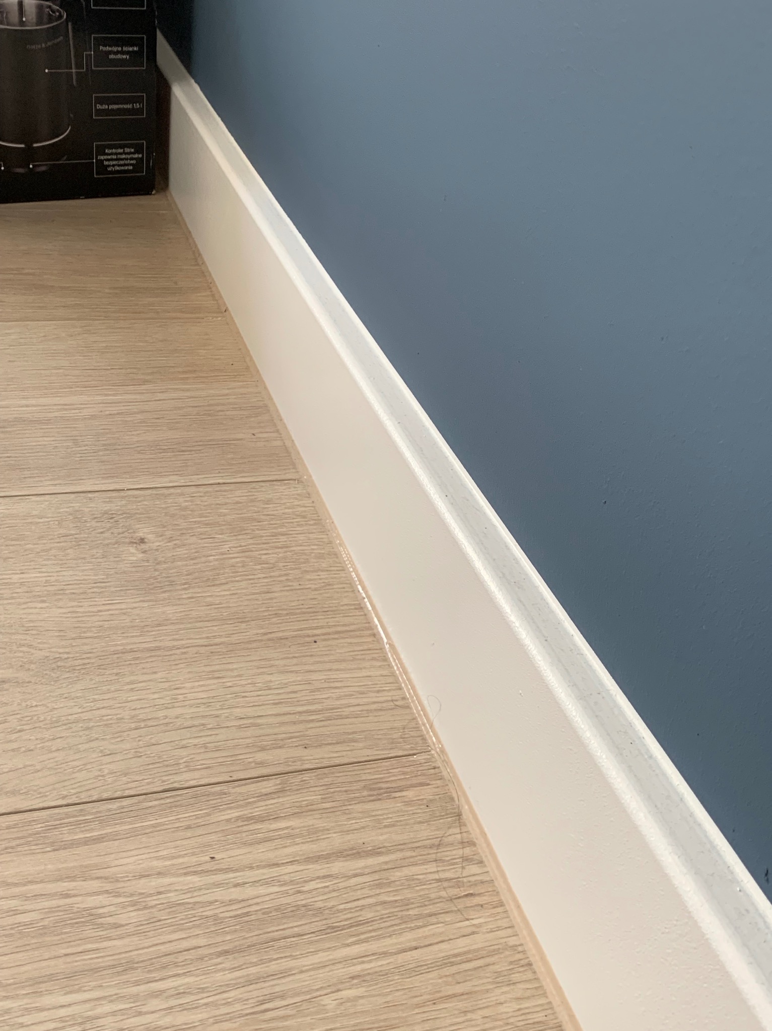 Biała listwa przypodłogowa na styku paneli podłogowych i ściany pomalowanej na niebiesko. Widoczne niedoskonałości malowania listwy. Fragment pudełka w lewym górnym rogu.