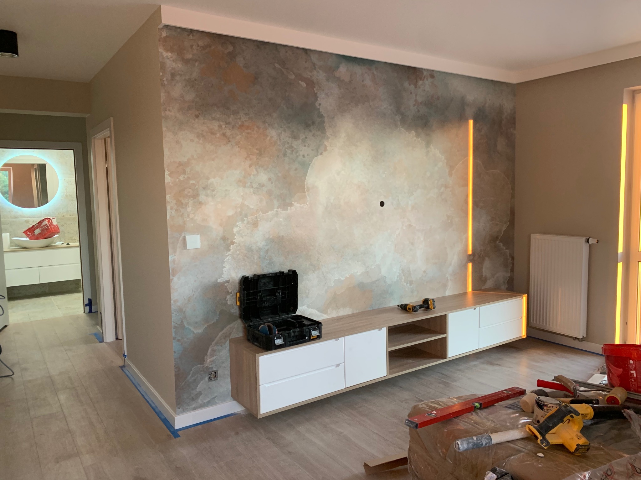 Wykończone wnętrze z dekoracyjną ścianą w odcieniach beżu i błękitu, podwieszana szafka RTV i narzędzia na podłodze. Widok na łazienkę z lustrem.