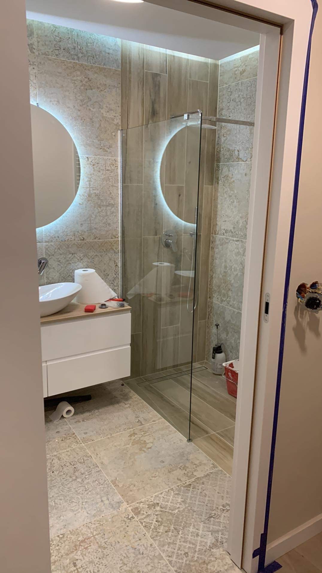 Wykończona łazienka z kabiną prysznicową typu walk-in, umywalką nablatową i okrągłymi lustrami z podświetleniem LED. Widoczne płytki w odcieniach beżu i szarości.