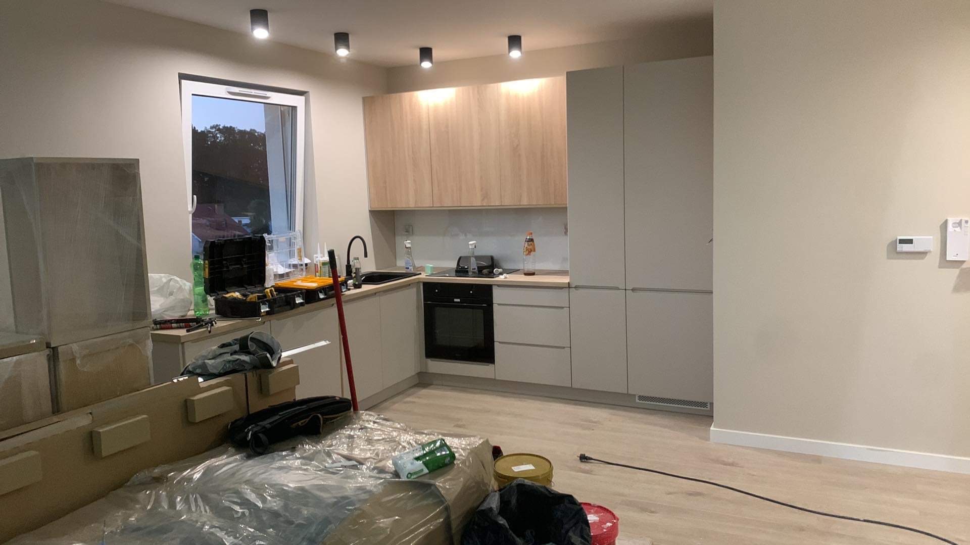 Jasna kuchnia w trakcie wykańczania: zabudowa meblowa, sprzęt AGD, narzędzia i folie ochronne na podłodze. Widok na okno i fragment zabudowy.