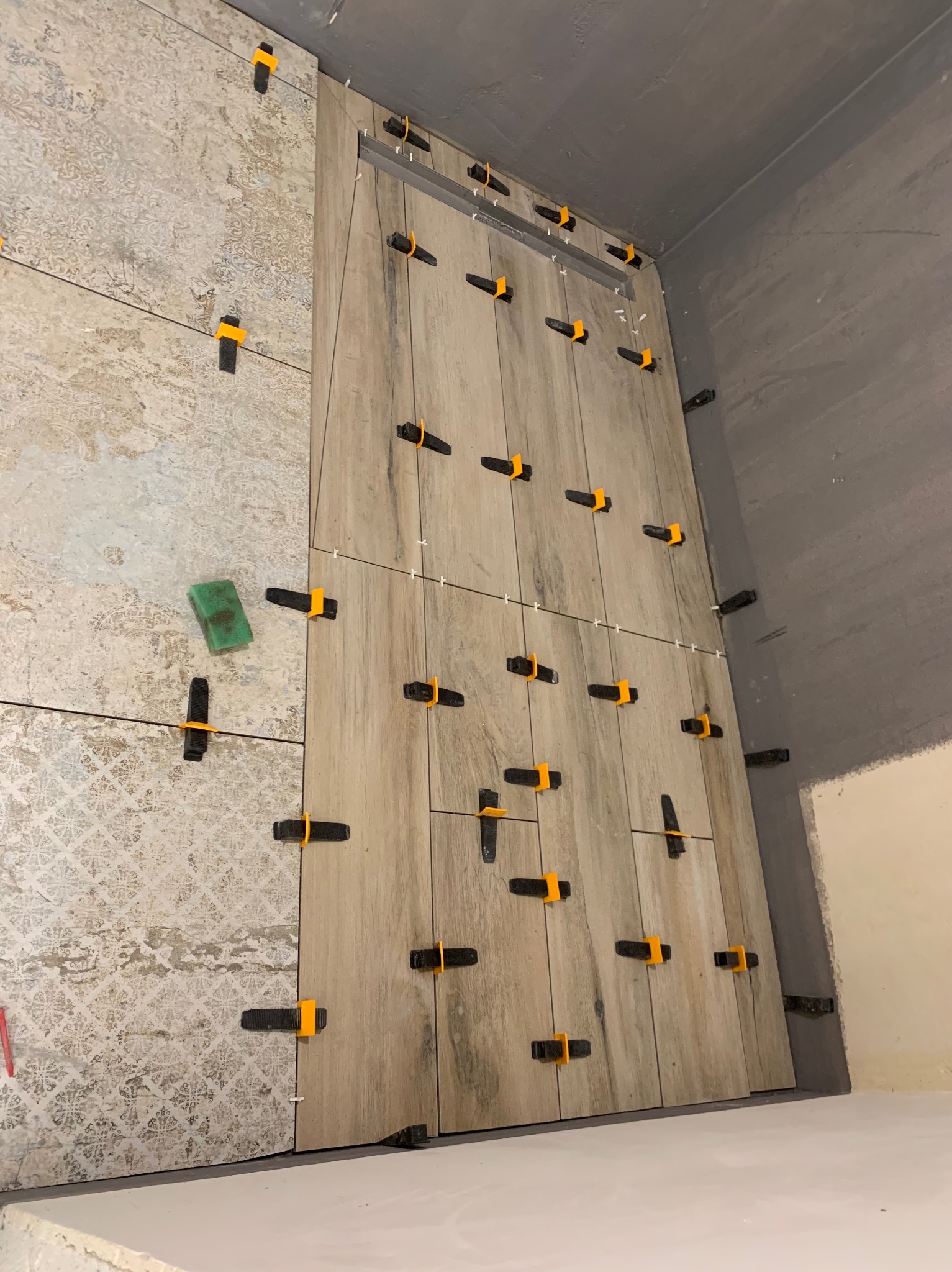 Układanie płytek imitujących drewno na ścianie z użyciem klipsów poziomujących. Widoczny fragment ściany z innym wzorem płytek i gąbka do czyszczenia.