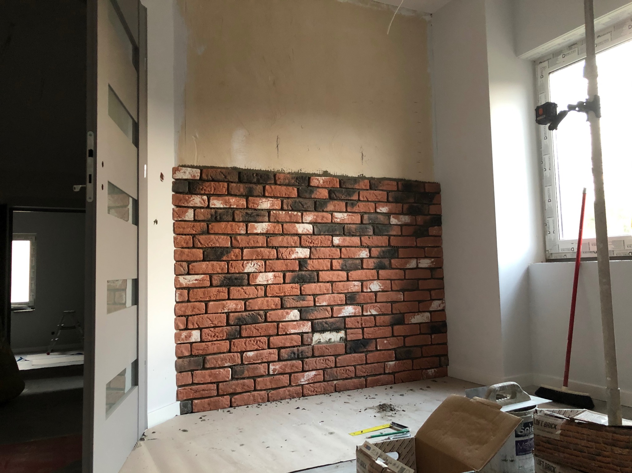 Ściana z cegły dekoracyjnej w trakcie montażu, widoczne narzędzia i materiały budowlane, laser krzyżowy przy oknie. Częściowo wykończone wnętrze.