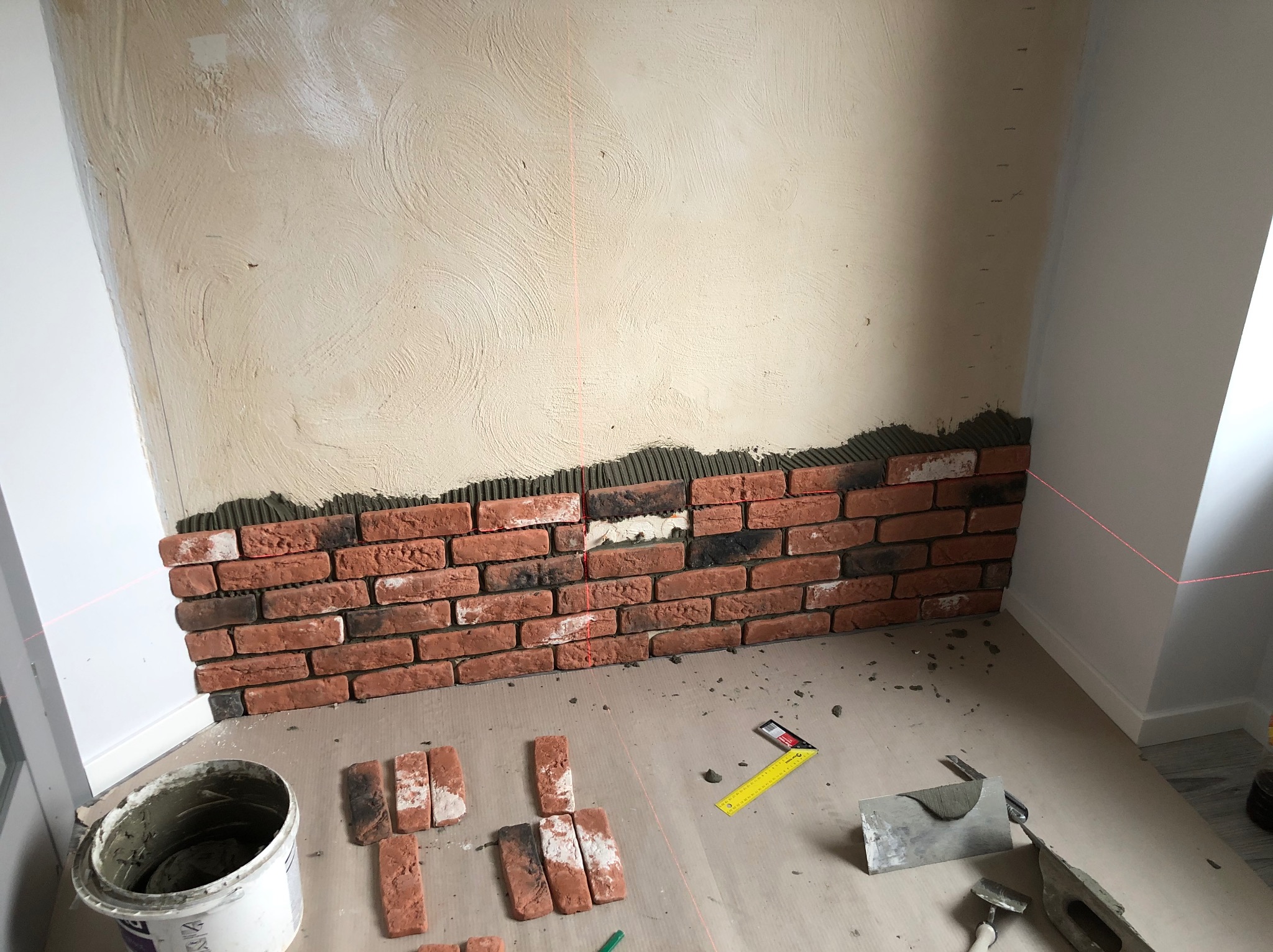 Montaż dekoracyjnej ściany z cegły: fragment ściany z ułożonymi cegłami, widoczny laserowy poziom, narzędzia glazurnicze i wiadro z zaprawą. Prace wykończeniowe w toku.