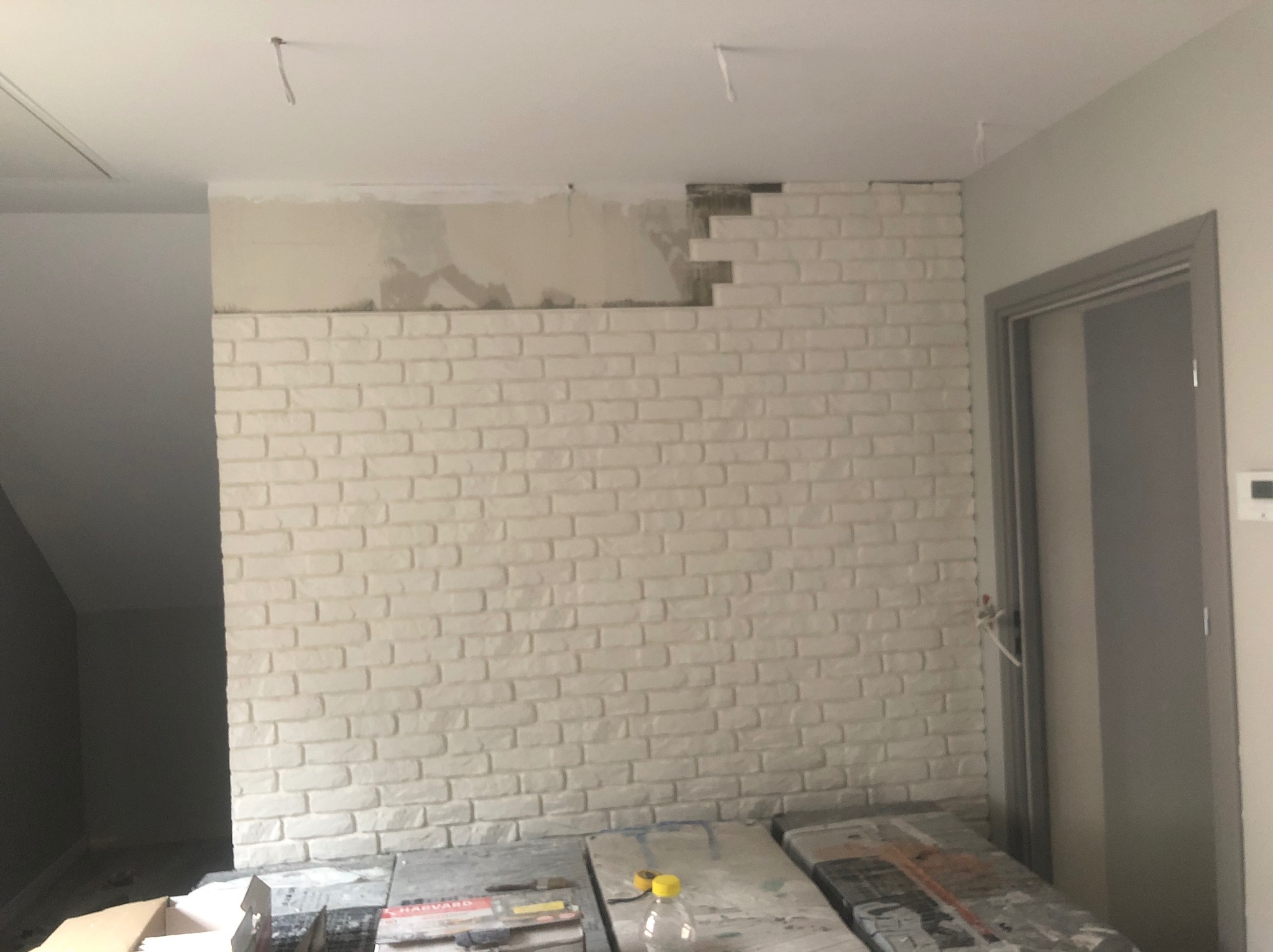 Ściana z białej cegły dekoracyjnej w trakcie montażu, widoczne niedokończone fragmenty i narzędzia pracy. Surowe wykończenie wnętrza, oświetlenie dzienne.