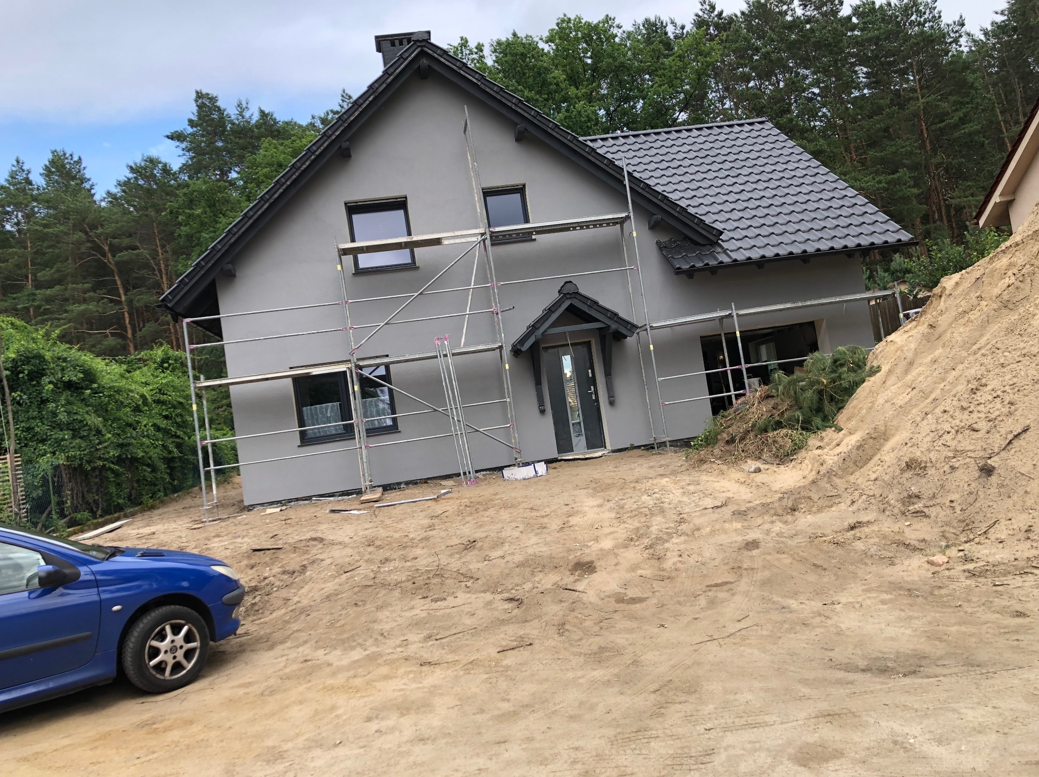 Szary dom jednorodzinny w trakcie malowania elewacji, z rusztowaniem i stertą piasku obok. Widoczny fragment niebieskiego samochodu.