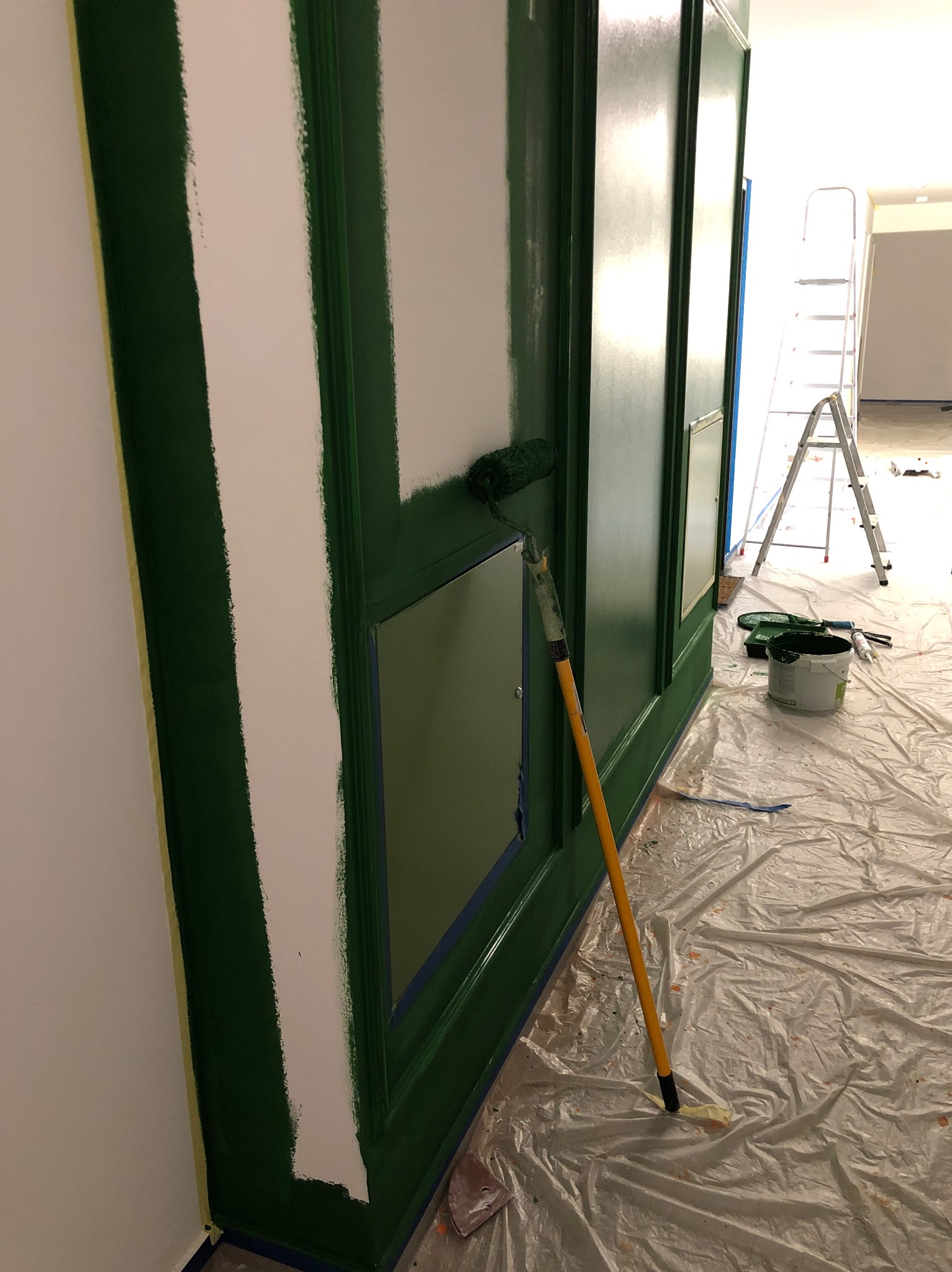 Malowanie dekoracyjnej ściany na zielono wałkiem. Widoczna drabina, wiadro z farbą i folia ochronna. Częściowo pomalowana ściana z białymi fragmentami.