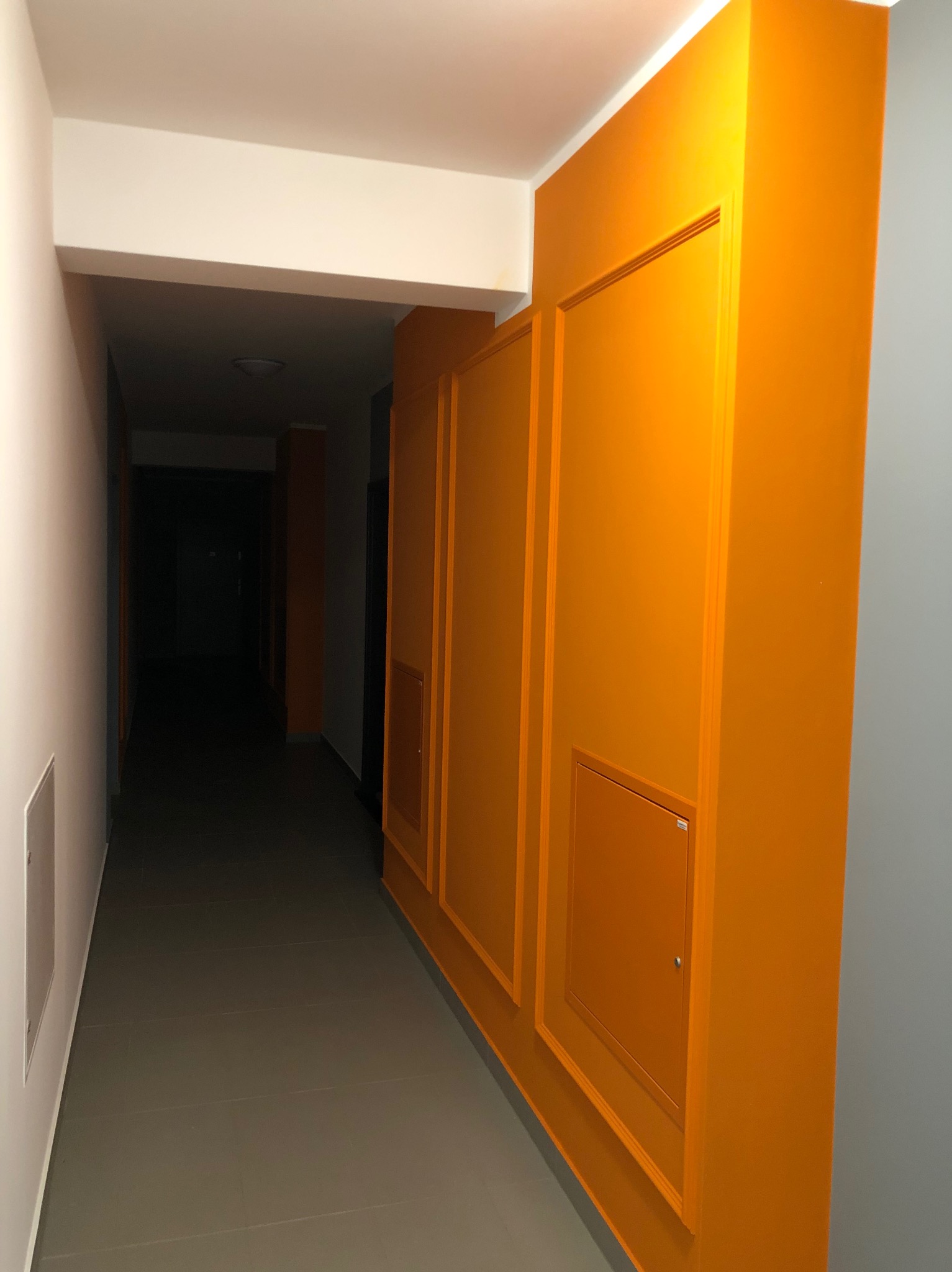 Korytarz po remoncie: pomarańczowa ściana z panelami i drzwiczkami rewizyjnymi, szara ściana i podłoga, biały sufit. Nowoczesne wykończenie wnętrza.