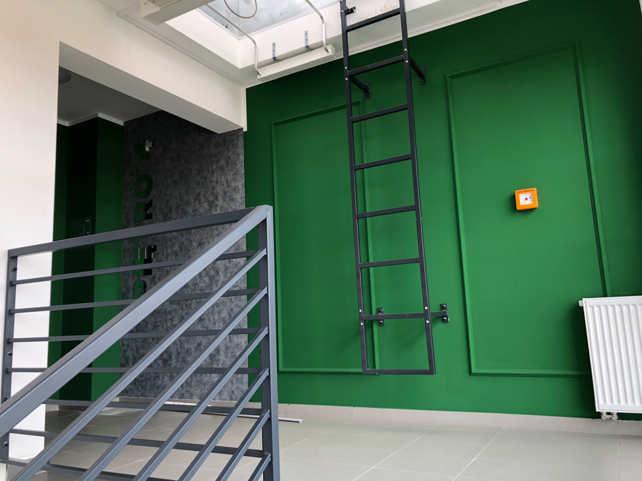 Nowoczesne wnętrze biurowe: zielona ściana z czarną drabinką, szara ściana z logo, biały grzejnik i szare schody z metalową barierką. Jasne płytki podłogowe.