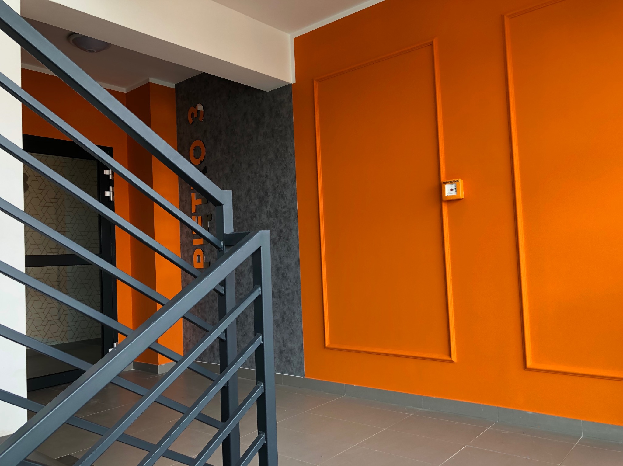 Klatka schodowa w Zielonej Górze po remoncie: pomarańczowe ściany z dekoracyjnymi ramami, szara ściana z numerem, szare metalowe barierki i płytki podłogowe.