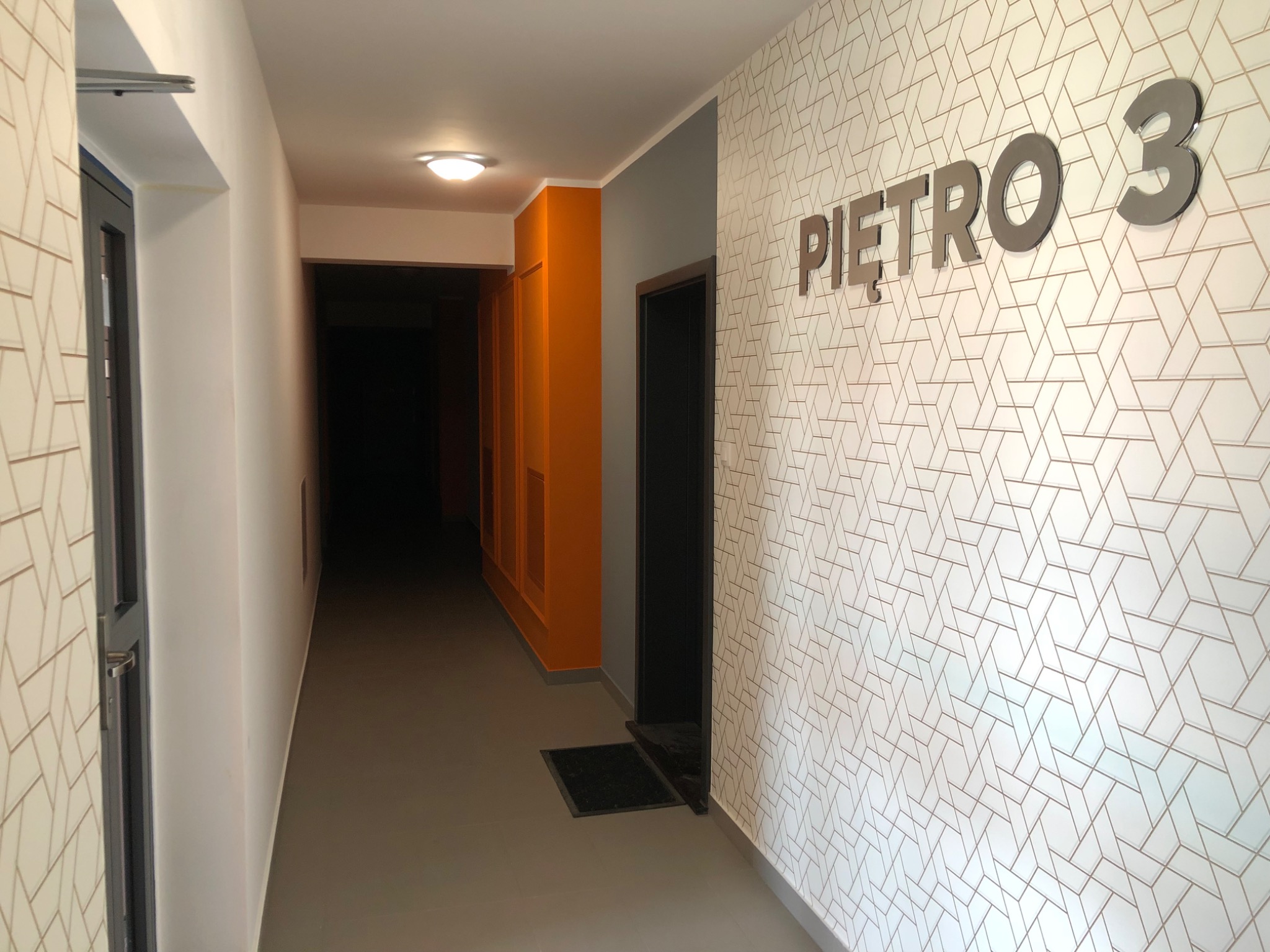Korytarz w budynku z geometryczną tapetą, napisem 'Piętro 3' i pomarańczową ścianą w tle. Widoczne drzwi i oświetlenie sufitowe.