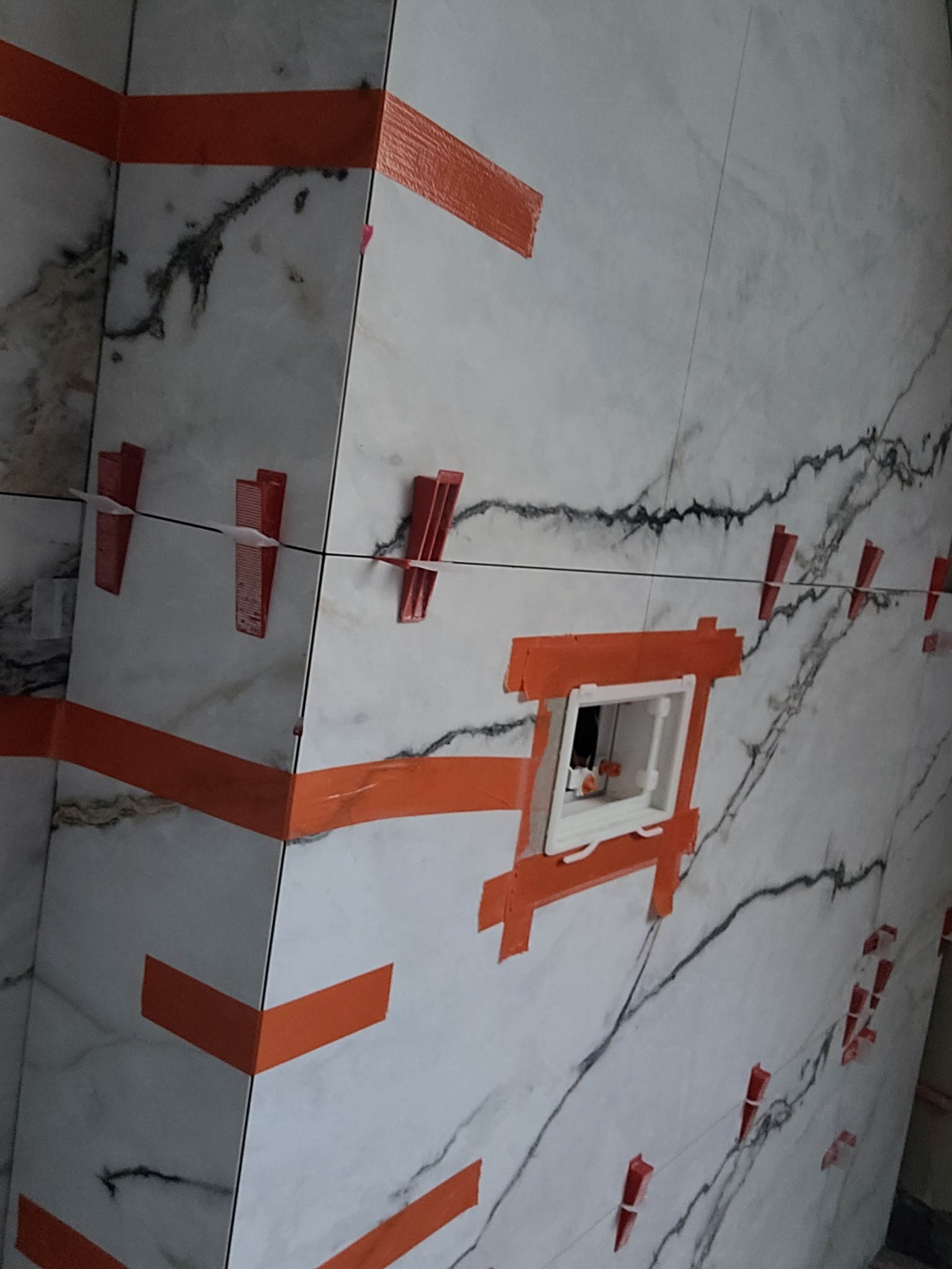 Układanie płytek imitujących marmur w łazience, z użyciem pomarańczowej taśmy maskującej i czerwonych klipsów poziomujących, widoczna zabudowa podtynkowego stelaża WC.