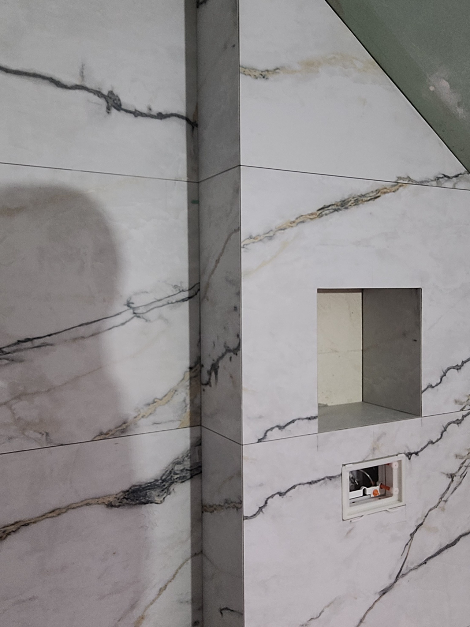 Ściana w łazience wyłożona białymi płytkami imitującymi marmur z wnęką i otworem na instalację hydrauliczną.