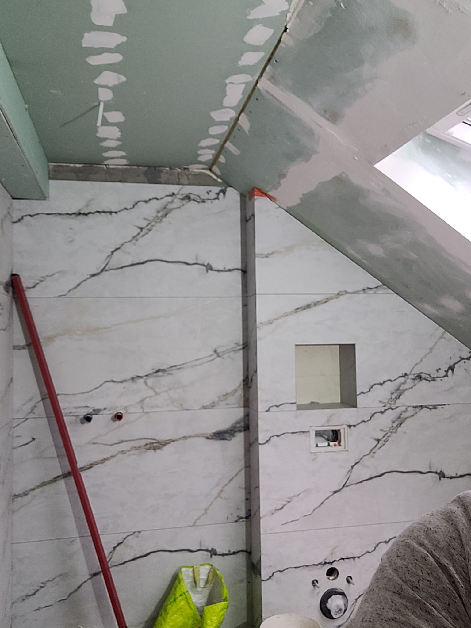 Montaż płytek imitujących marmur na ścianie skośnej łazienki na poddaszu, widoczne otwory na armaturę, zielona torba budowlana i częściowo zagruntowany sufit z płyt kartonowo-gipsowych.