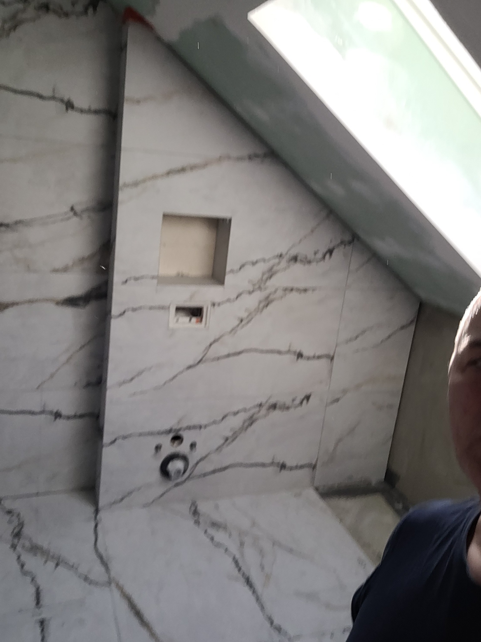 Ściana w łazience z ukośnym sufitem, wyłożona białymi płytkami imitującymi marmur z ciemnymi żyłami, widoczne otwory na armaturę i wnęka w ścianie, częściowo widoczna osoba.