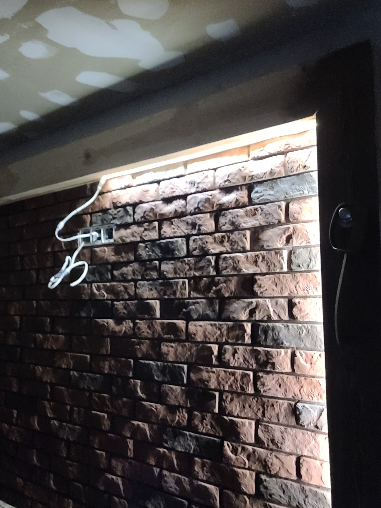 Ściana wyłożona dekoracyjną cegłą w trakcie montażu, widoczne kable elektryczne wystające ze ściany, sufit w trakcie wykańczania płytami gipsowo-kartonowymi.