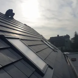 Montaż okien dachowych na skośnym dachu pokrytym ciemną dachówką ceramiczną, widoczna sylwetka dekarza w górnym rogu oraz fragment zabudowy w tle.