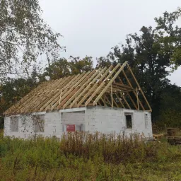 Wznoszenie więźby dachowej na surowym budynku z betonu komórkowego, widok na konstrukcję z drewnianych belek na tle jesiennego krajobrazu.