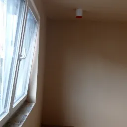 Wnętrze pokoju po świeżym nałożeniu gładzi, widoczne gładkie ściany w neutralnym odcieniu beżu, okno z białymi ramami i wystający biały, cylindryczny punkt oświetleniowy na suficie.