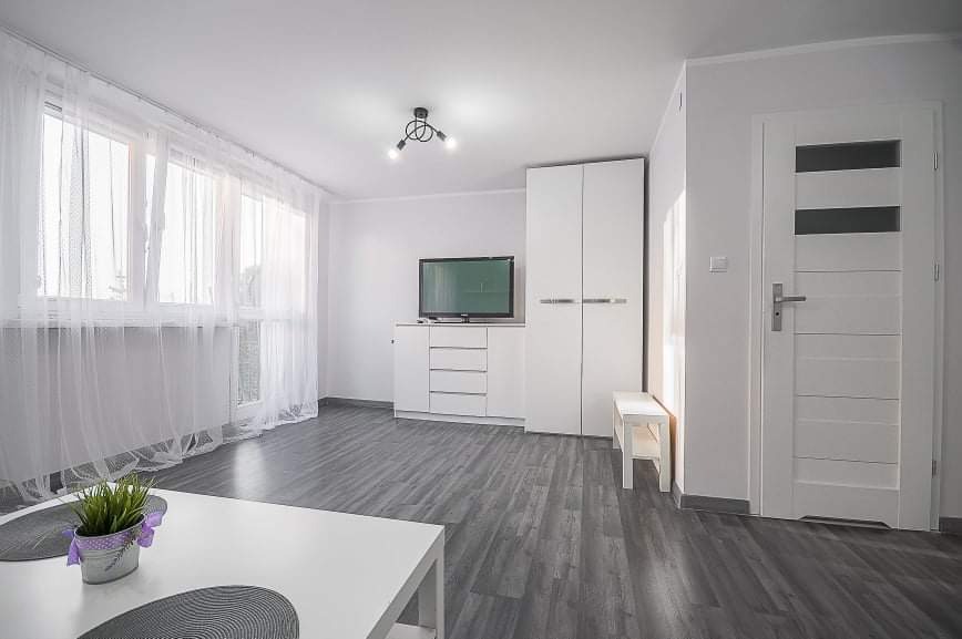 Jasne, minimalistyczne wnętrze pokoju z szarymi panelami podłogowymi, białymi ścianami, szafą, komodą z telewizorem, oknem z firankami oraz białymi drzwiami z przeszkleniami.