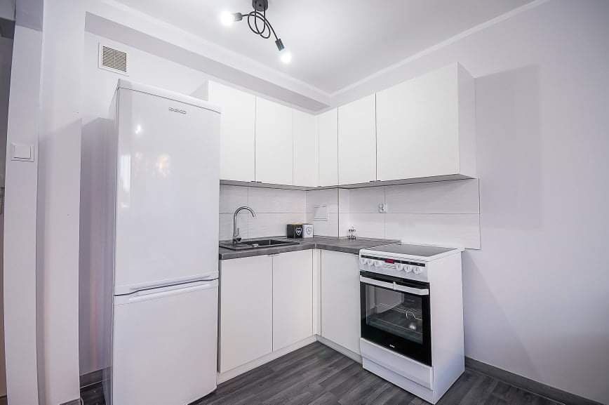 Jasna kuchnia w nowoczesnym stylu z białymi szafkami, szarym blatem, lodówką i piekarnikiem, oświetlona lampą sufitową z trzema punktami świetlnymi.