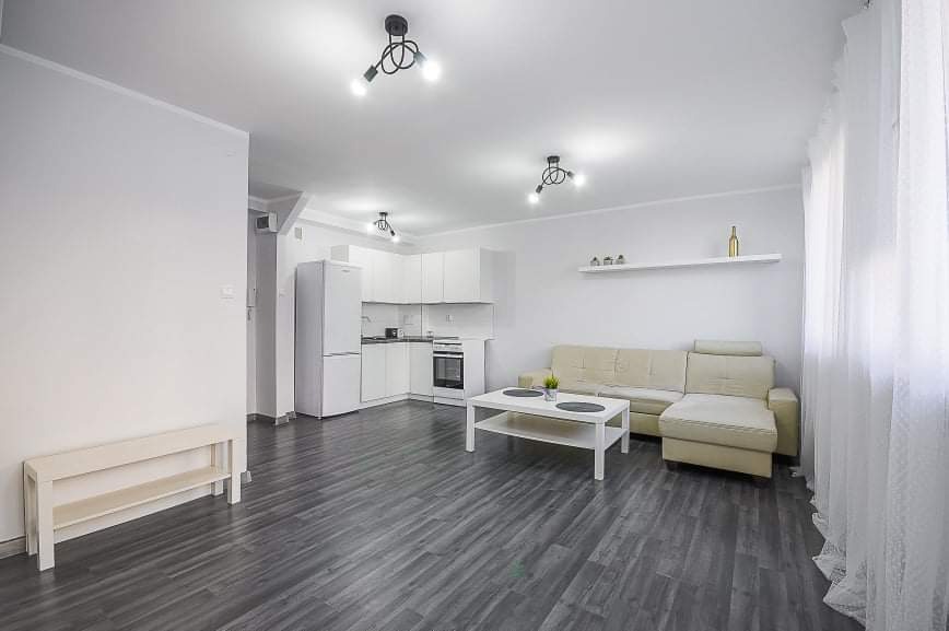 Jasne, nowocześnie wykończone wnętrze mieszkania z szarymi panelami podłogowymi, białymi ścianami, aneksem kuchennym i beżową sofą narożną. Na ścianie półka z dekoracjami i butelką. Okno zasłonięte...