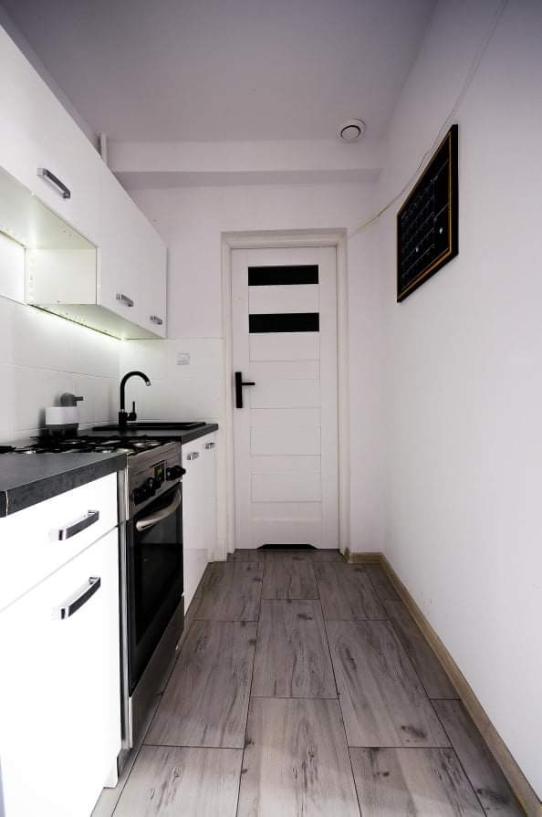 Wąska kuchnia z białymi szafkami, czarnym blatem i drewnopodobną podłogą, widok na drzwi z czarnymi poziomymi paskami i tablicę na ścianie.