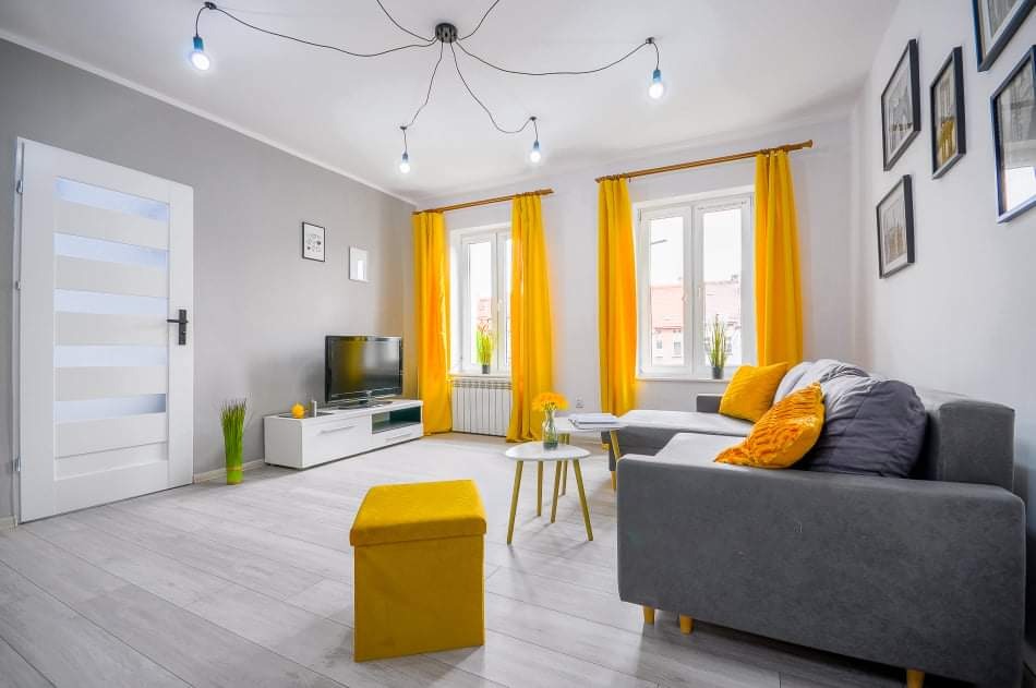 Jasne wnętrze salonu z szarą sofą narożną, żółtymi zasłonami i pufem, białymi drzwiami z przeszkleniami i lampami wiszącymi na kablach.