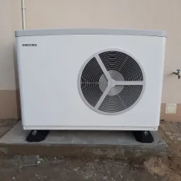 Biała pompa ciepła Stiebel Eltron zamontowana na betonowej podstawie przed budynkiem z beżową elewacją, widoczny kran na ścianie i fragment chodnika z ziemią.