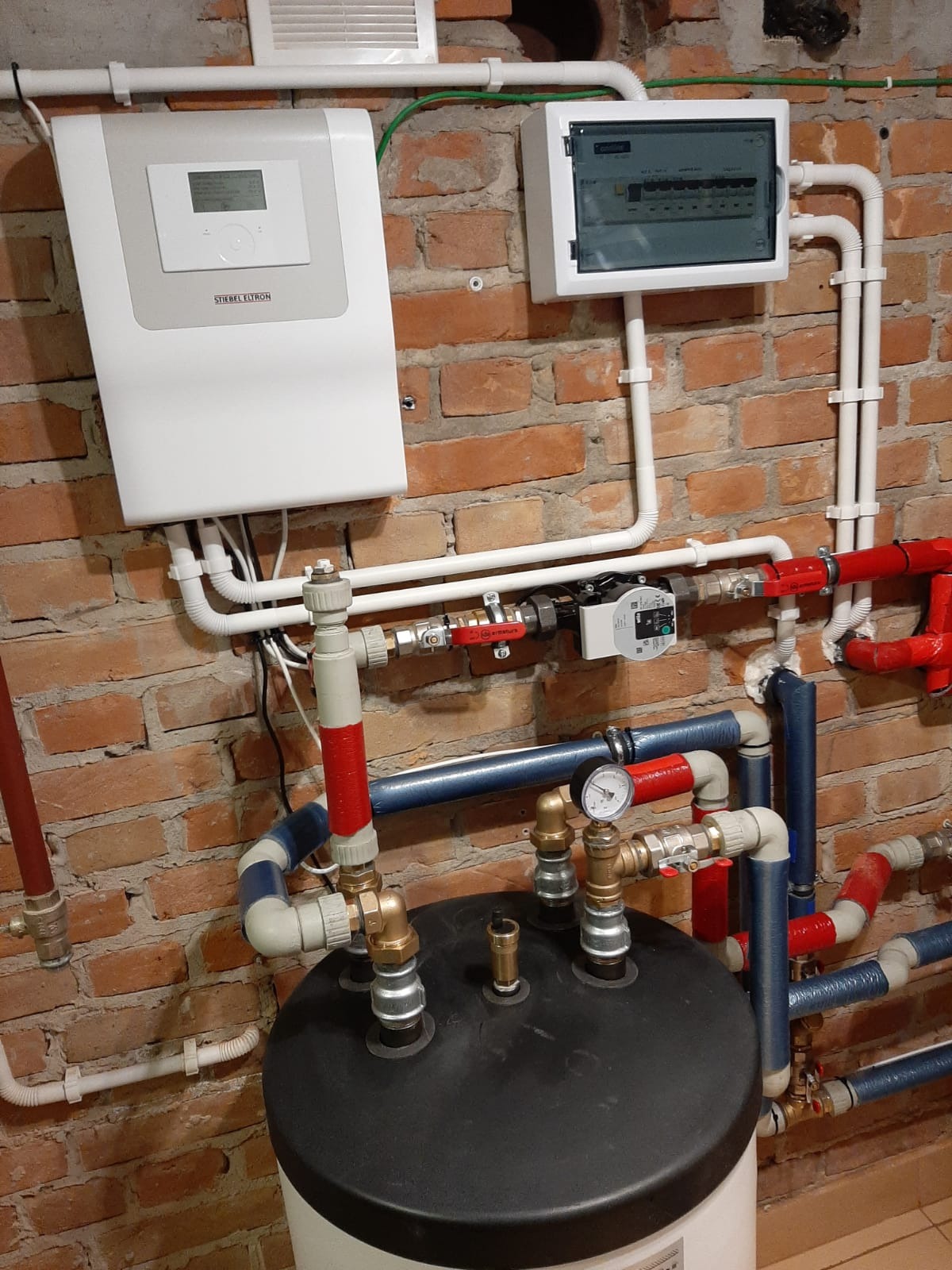 Instalacja z kotłem i rozdzielnicą elektryczną na tle ceglanej ściany, widoczne rury w kolorach białym, czerwonym i niebieskim, manometr oraz biały panel sterowania Stiebel Eltron.