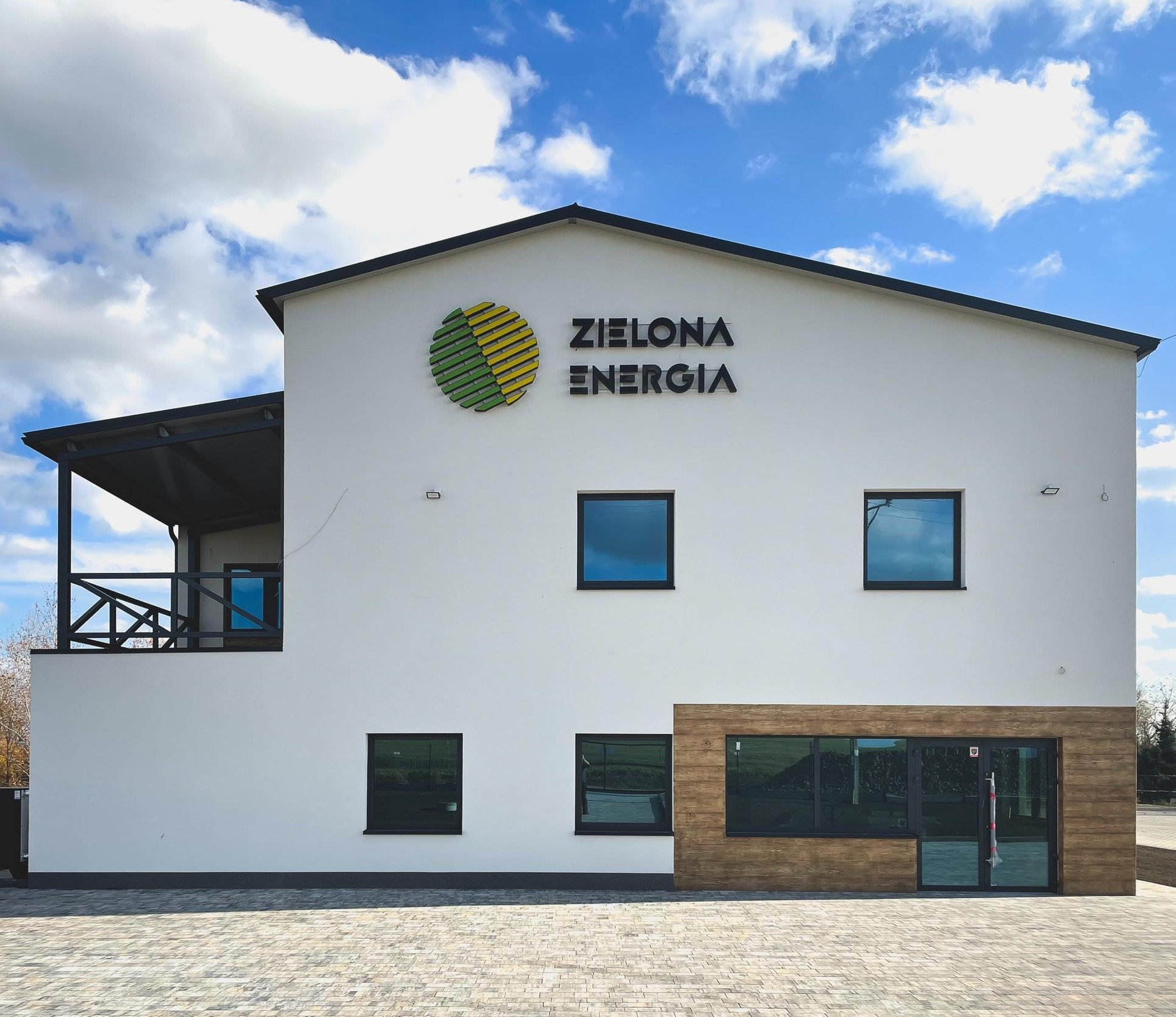 Nowoczesny budynek z białymi ścianami, z logo firmy 'Zielona Energia' i oknami odbijającymi błękitne niebo, wykończony drewnianymi panelami.