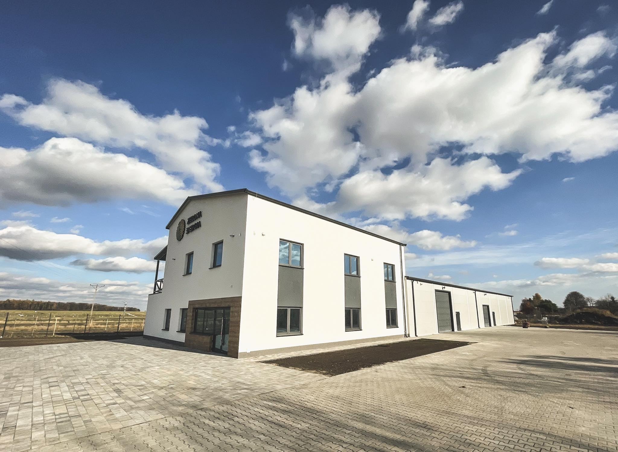 Nowoczesny budynek biurowy z białymi ścianami, szarymi oknami i kostką brukową na placu przed nim, błękitne niebo z chmurami w tle.