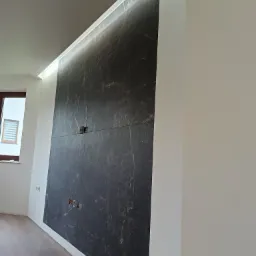 Ściana wykończona płytami imitującymi czarny marmur z widocznymi gniazdkami i włącznikami, podświetlona listwą LED przy suficie, widoczny fragment okna i drewniana podłoga.