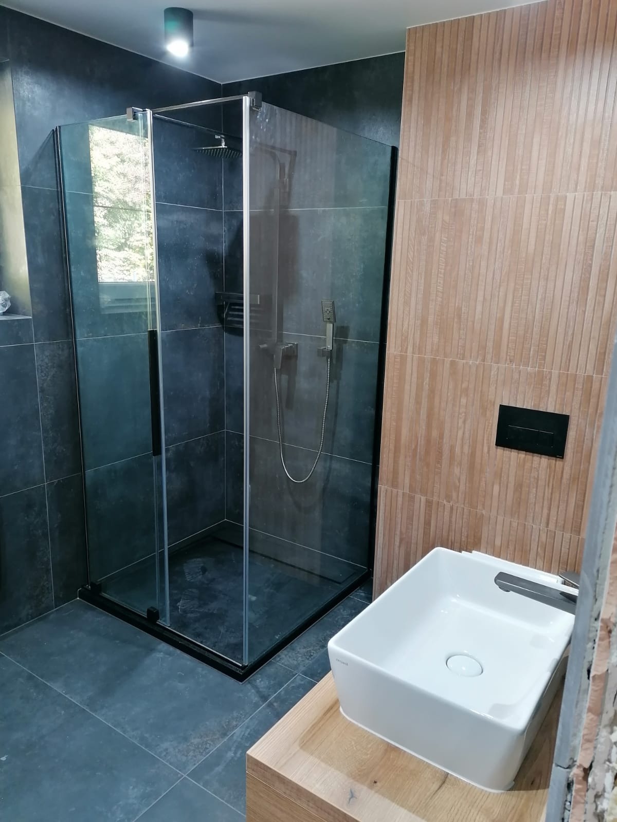 Nowoczesna łazienka z kabiną prysznicową typu walk-in z czarnymi profilami, umywalką nablatową na drewnianym blacie i płytkami imitującymi drewno na ścianie.