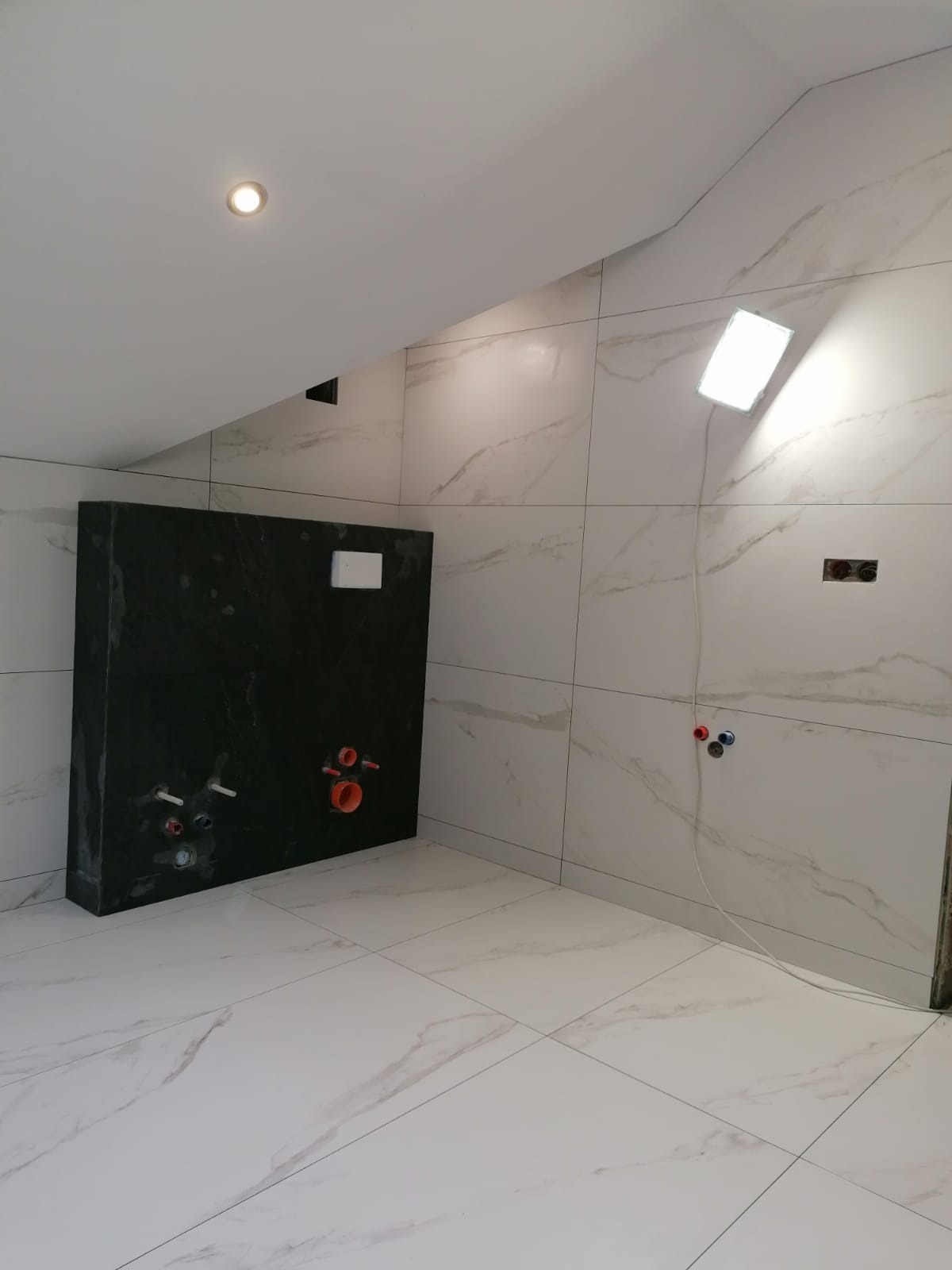 Nowoczesna łazienka w trakcie wykańczania, widoczne białe płytki imitujące marmur na ścianach i podłodze, czarny zabudowany stelaż podtynkowy WC z instalacją hydrauliczną, oświetlenie robocze.