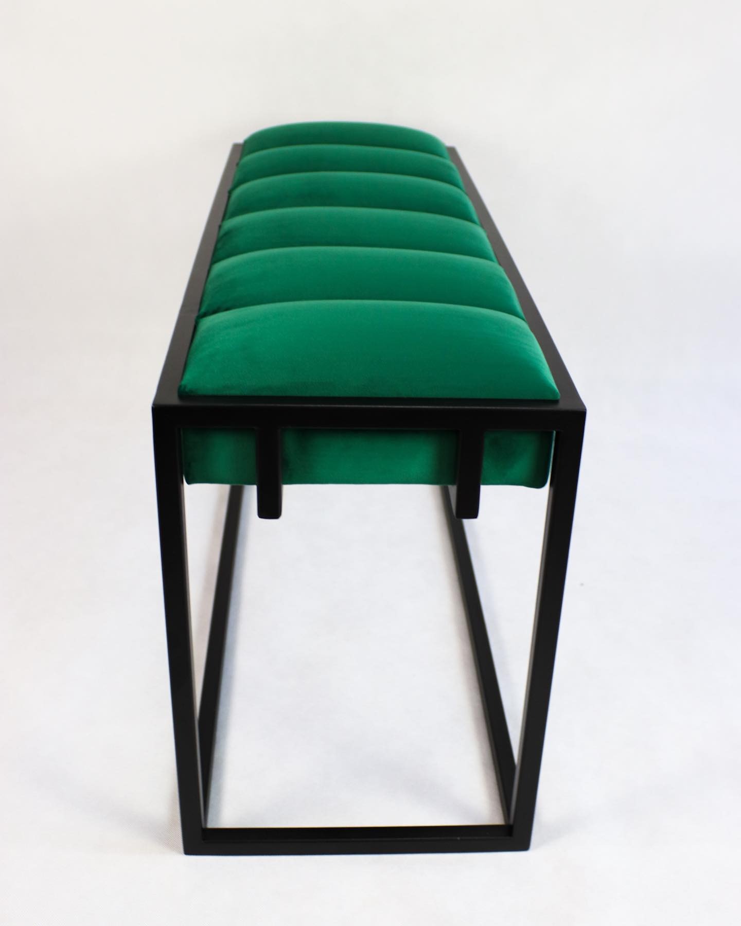 Nowoczesna ławka z czarną metalową ramą i zielonym welurowym siedziskiem z podziałem na segmenty, na białym tle.