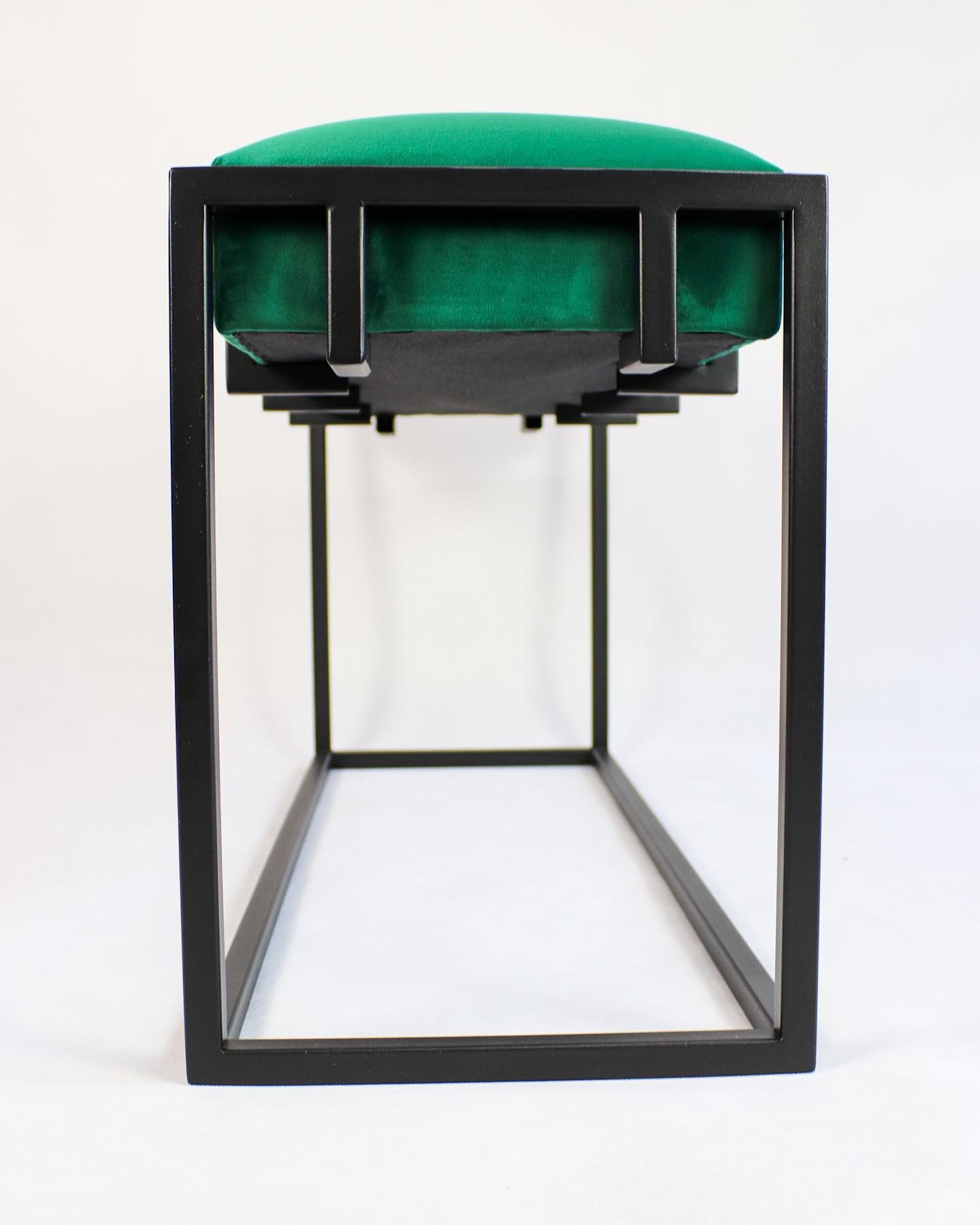 Niski stołek z zielonym, welurowym siedziskiem i czarną, metalową ramą o geometrycznym kształcie na białym tle.