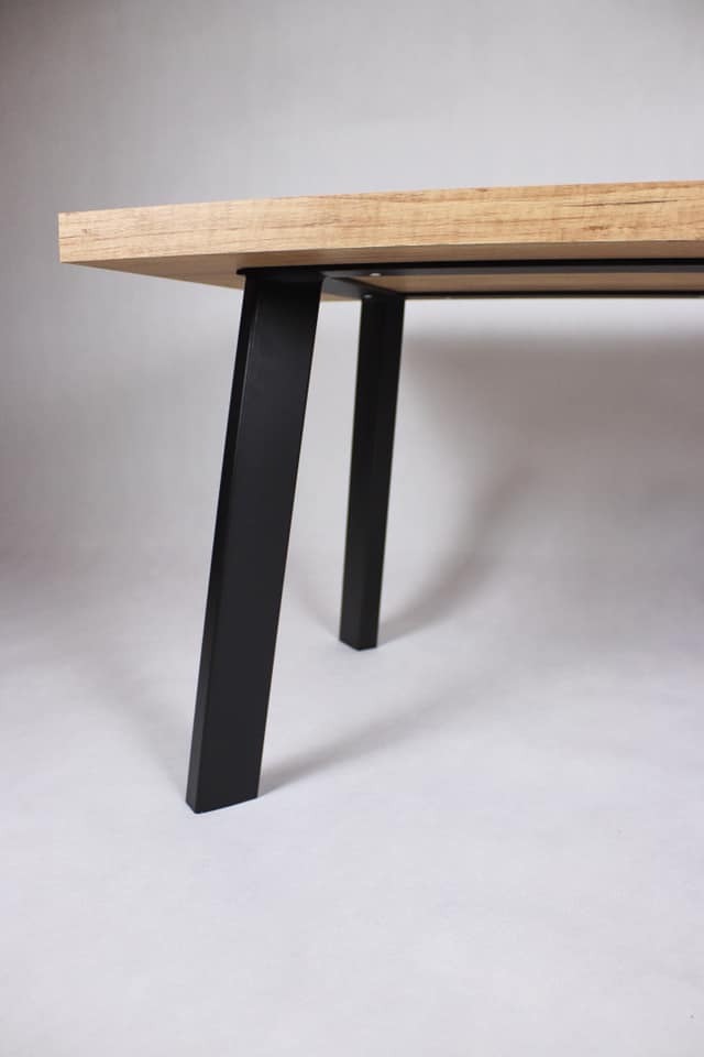 Fragment stołu z drewnianym blatem i czarnymi, metalowymi nogami o kwadratowym przekroju, na jasnym tle.