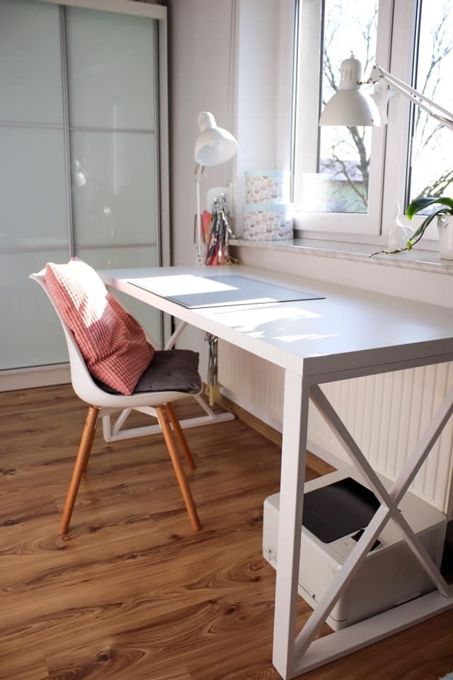 Białe biurko na wymiar z blatem z płyty laminowanej, oparte na konstrukcji z profili stalowych, umieszczone w jasnym pokoju obok okna i szafy przesuwnej, z krzesłem i drukarką pod spodem.