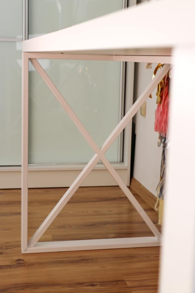 Biały stolik z metalową konstrukcją w kształcie litery X, widoczne dekoracje wiszące w tle, na podłodze panele.