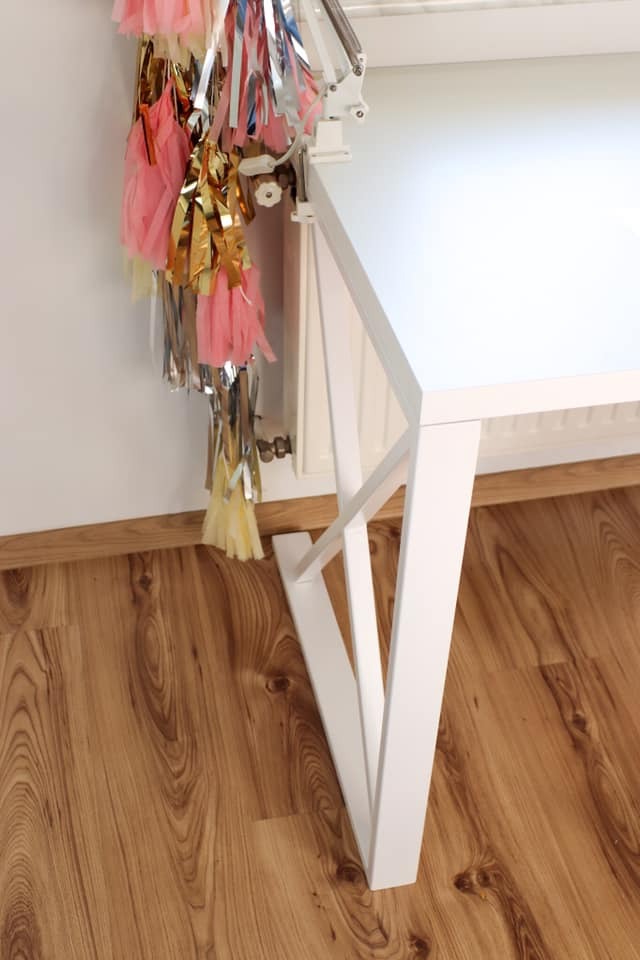 Białe biurko na tle drewnianej podłogi, z dekoracjami w odcieniach różu, złota i srebra przymocowanymi do blatu za pomocą białego uchwytu.