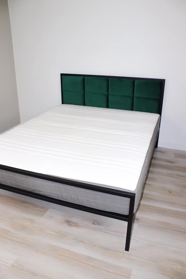 Łóżko na metalowej ramie malowanej proszkowo na czarno, z wezgłowiem obitym zielonym welurem, ustawione na podłodze imitującej drewno.