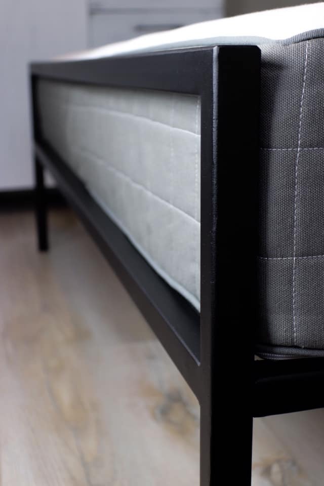 Metalowa rama łóżka z szarym materacem, widok z niskiego kąta, ukazujący detale ramy i tekstury materaca.